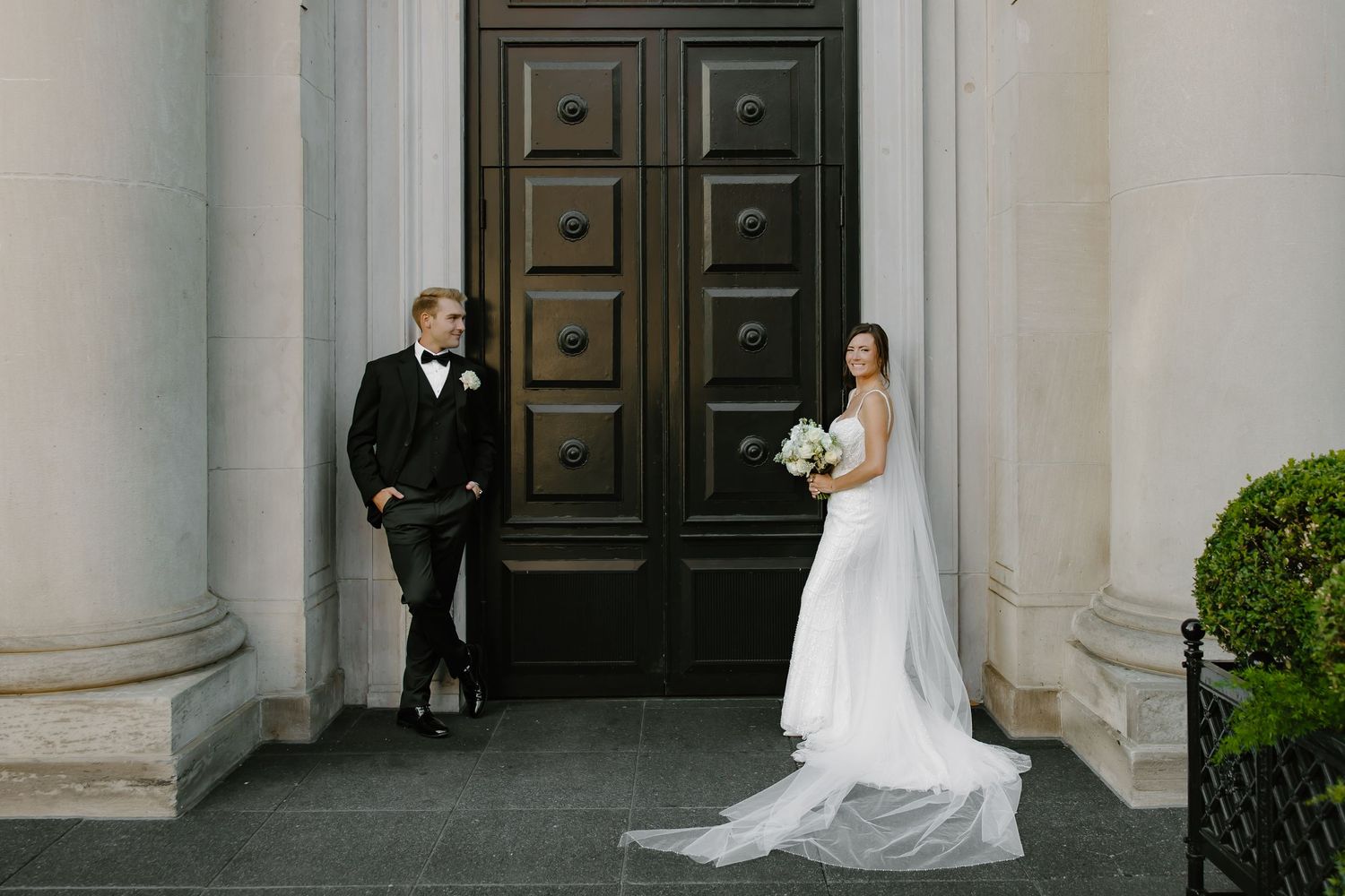 Wedding couple poses separately against grand wooden doors with classical columns.