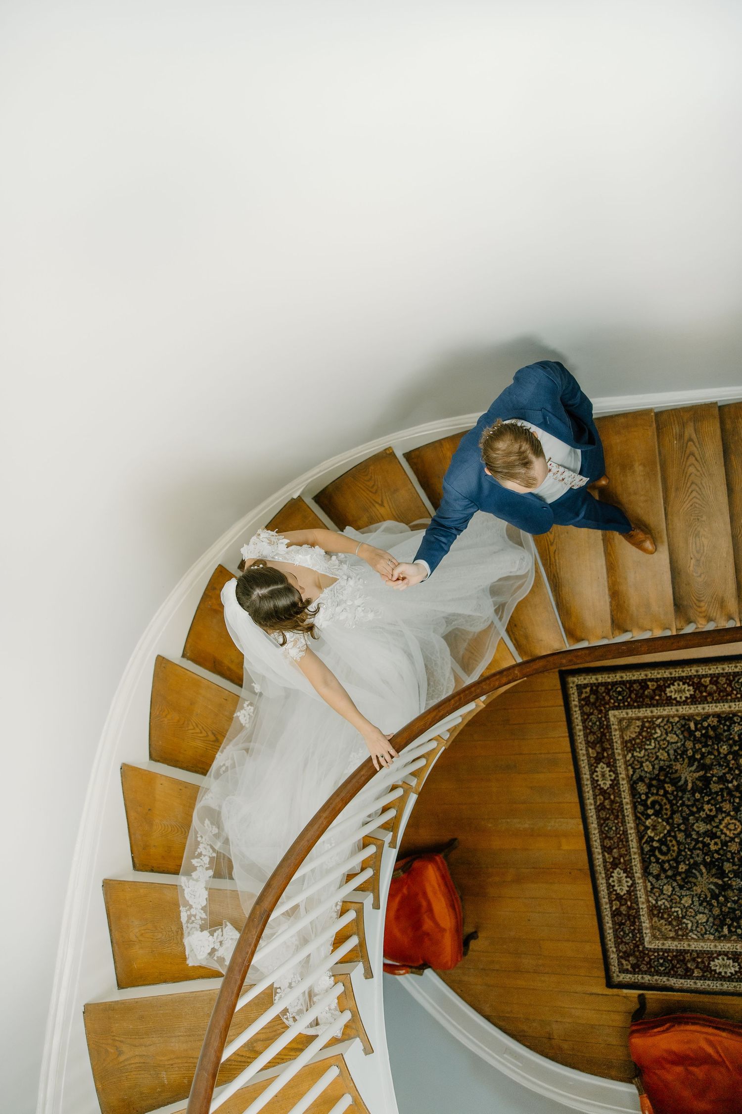 Overhead view of a bride in white dress descending curved wooden staircase.