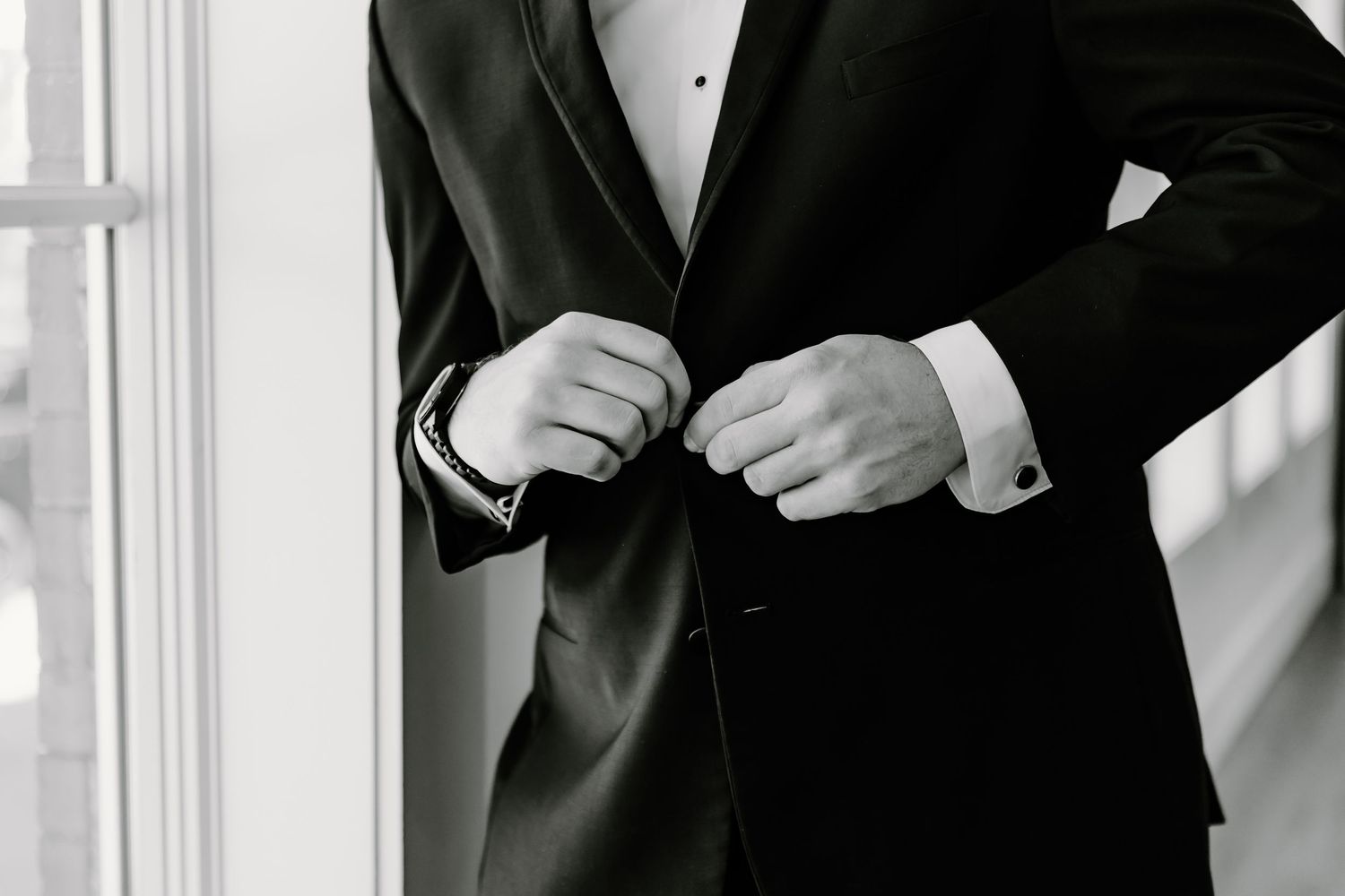 Men in black tuxedos preparing for formal event near windows.