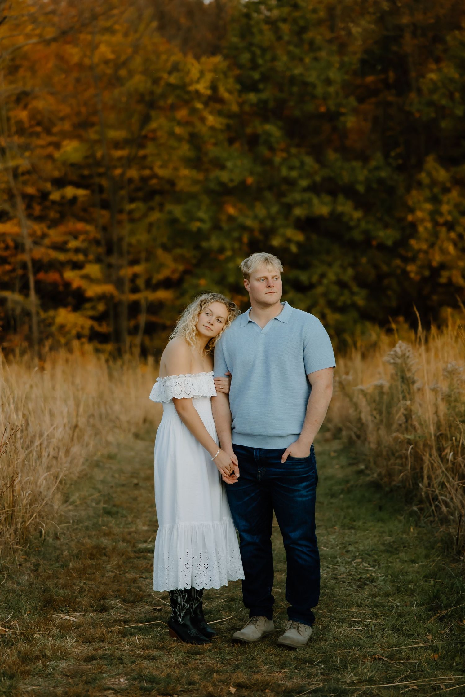 Couple poses together on a path with autumn foliage in the background during golden hour.