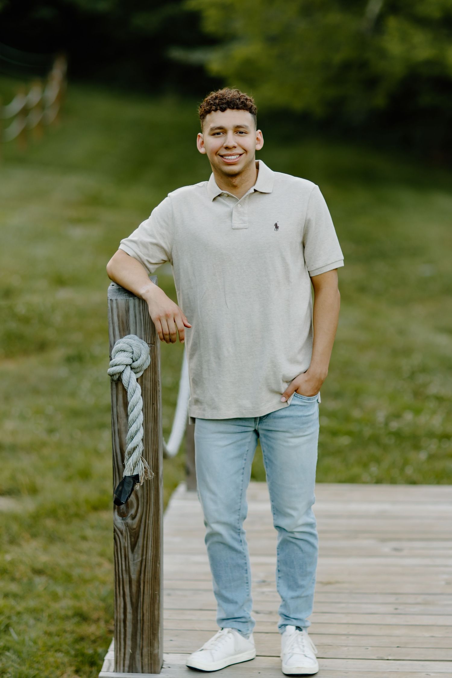 A person in a light gray polo shirt and light blue jeans stands casually on a wooden deck surrounded by greenery.