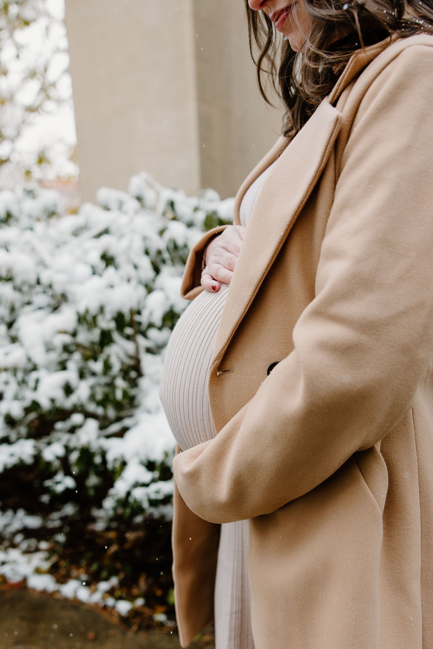 Close-up of pregnant belly in cream dress with camel coat against snowy winter background.