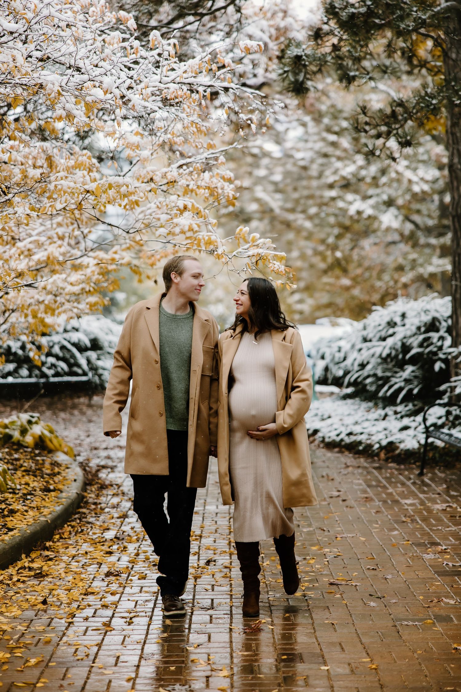 Couple walking on brick pathway surrounded by snow-covered autumn trees.