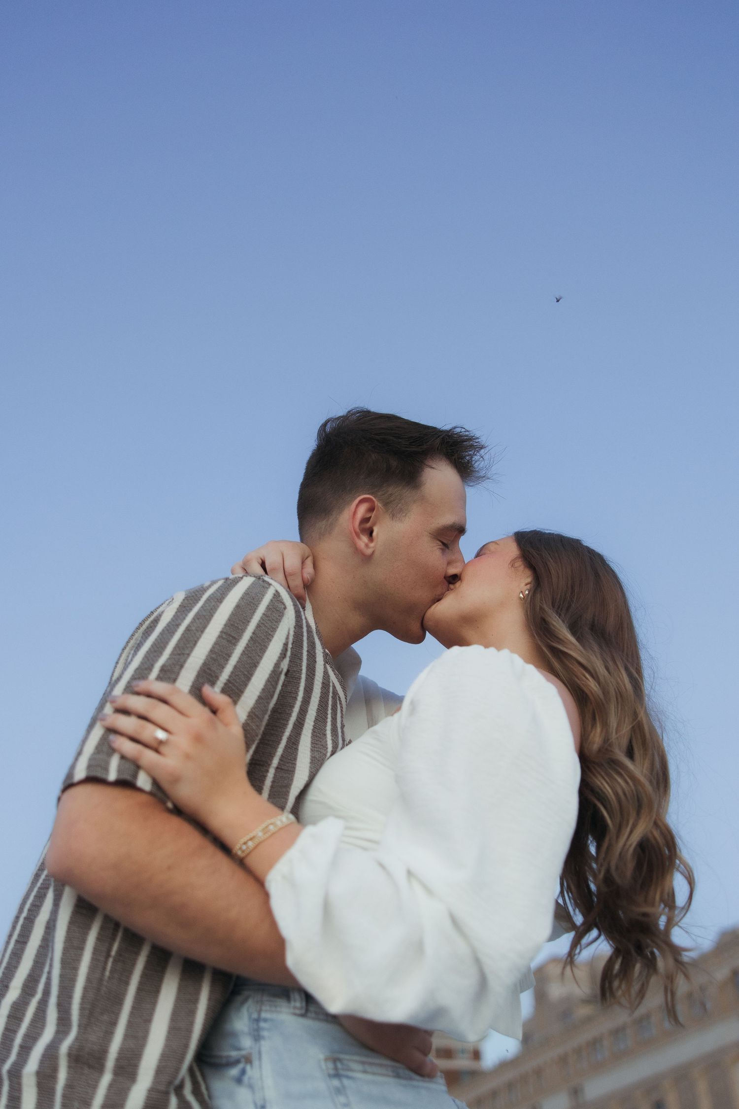 A couple shares a romantic kiss outdoors against a clear blue sky, embracing each other tenderly.