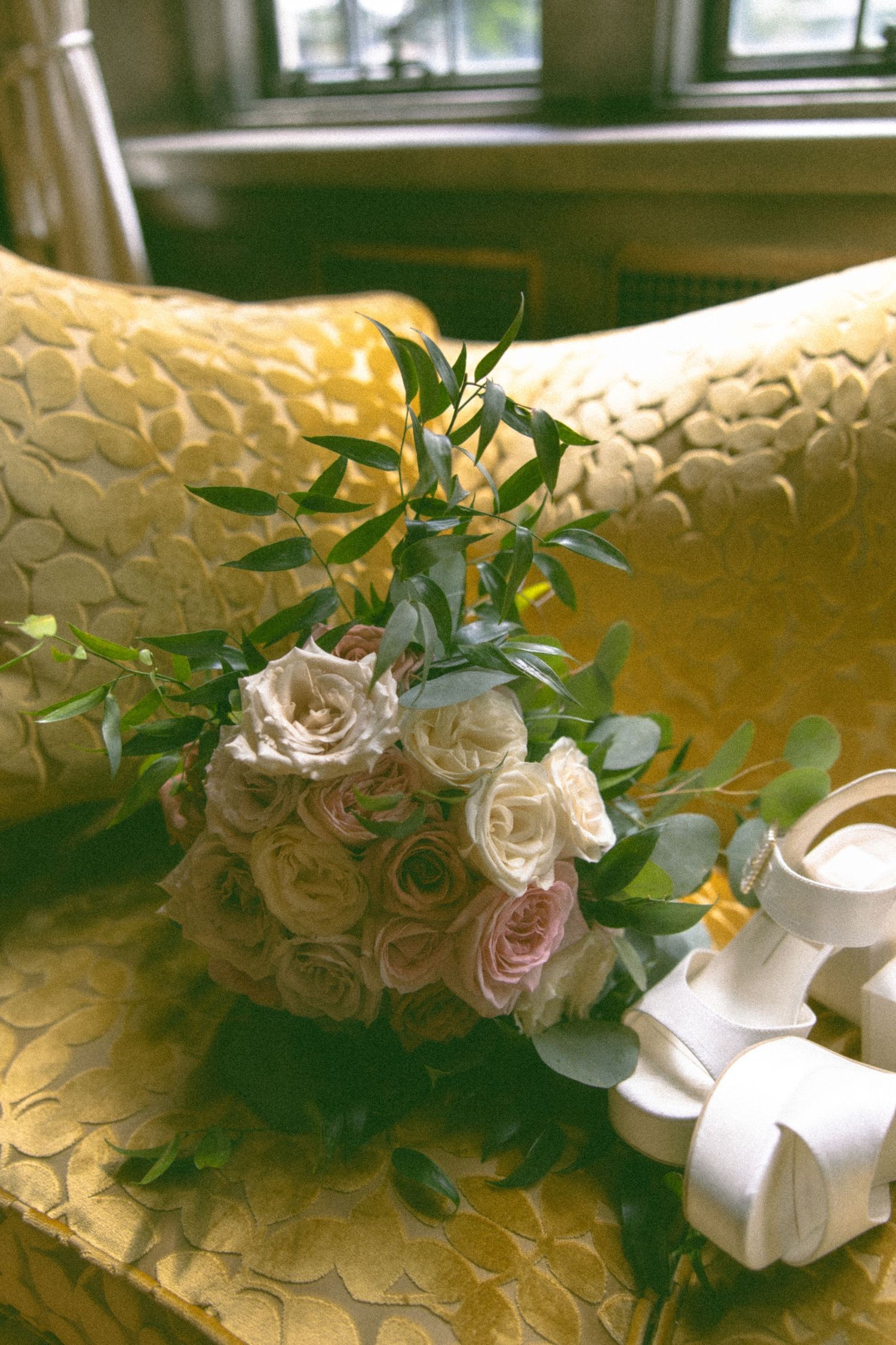 Delicate white and pink roses with greenery arranged on textured gold surface near window.