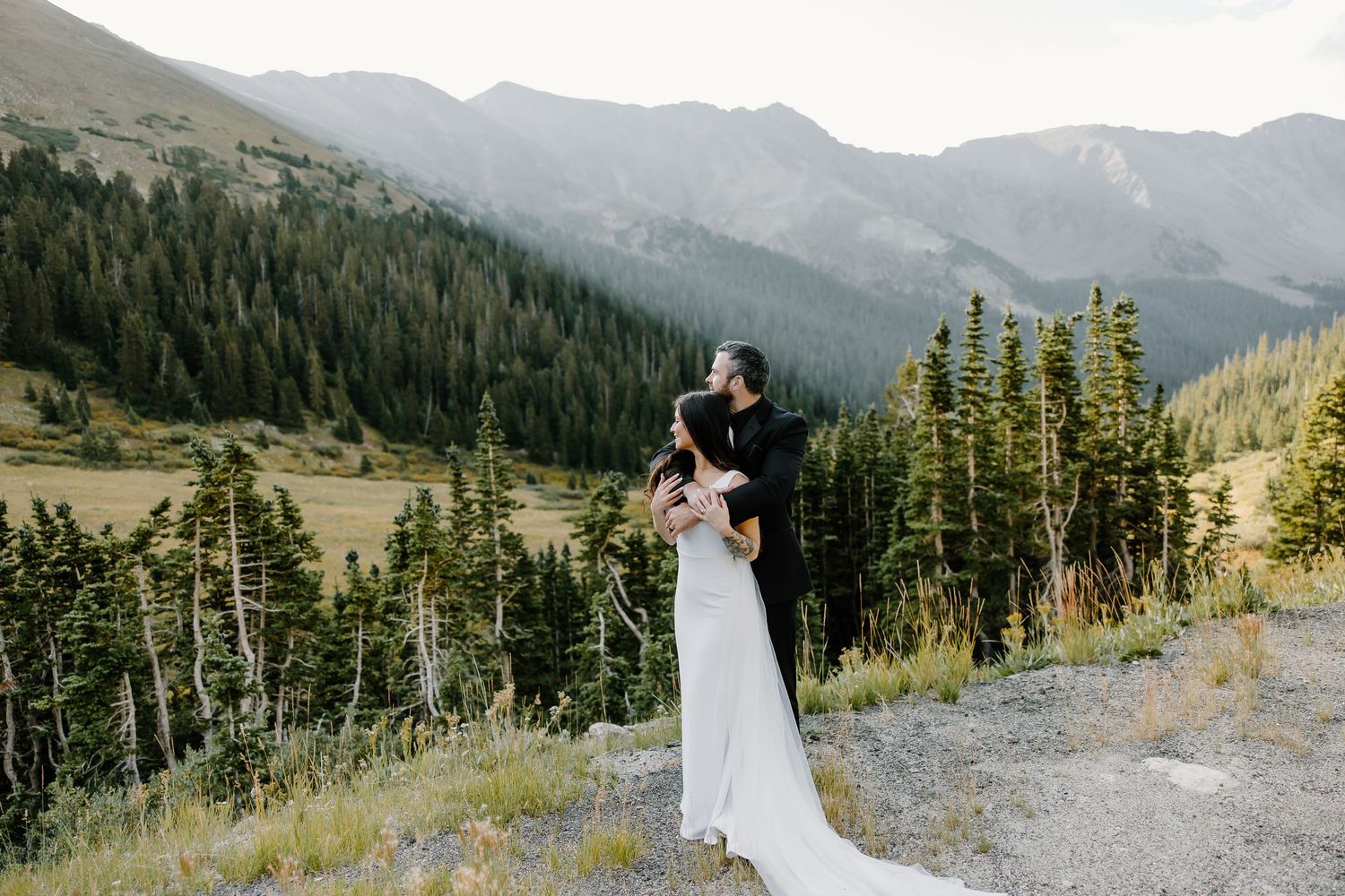 Wedding couple standing together against a scenic mountain backdrop with evergreen trees.