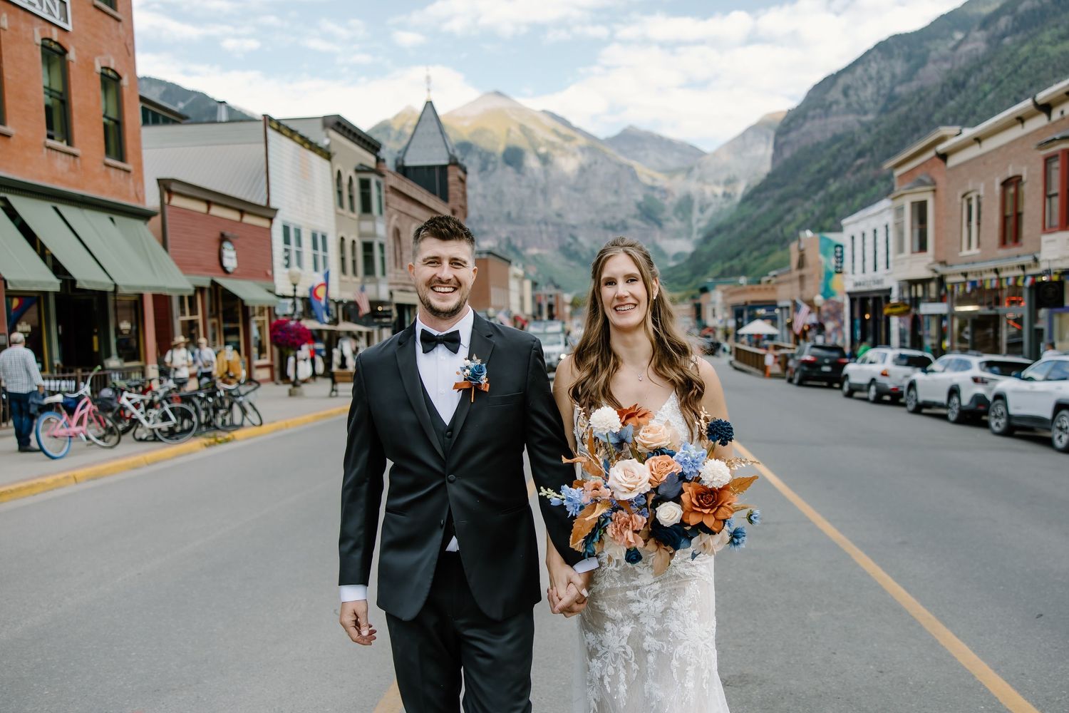 Newlyweds smile while walking down a historic mountain town's main street with colorful buildings.
