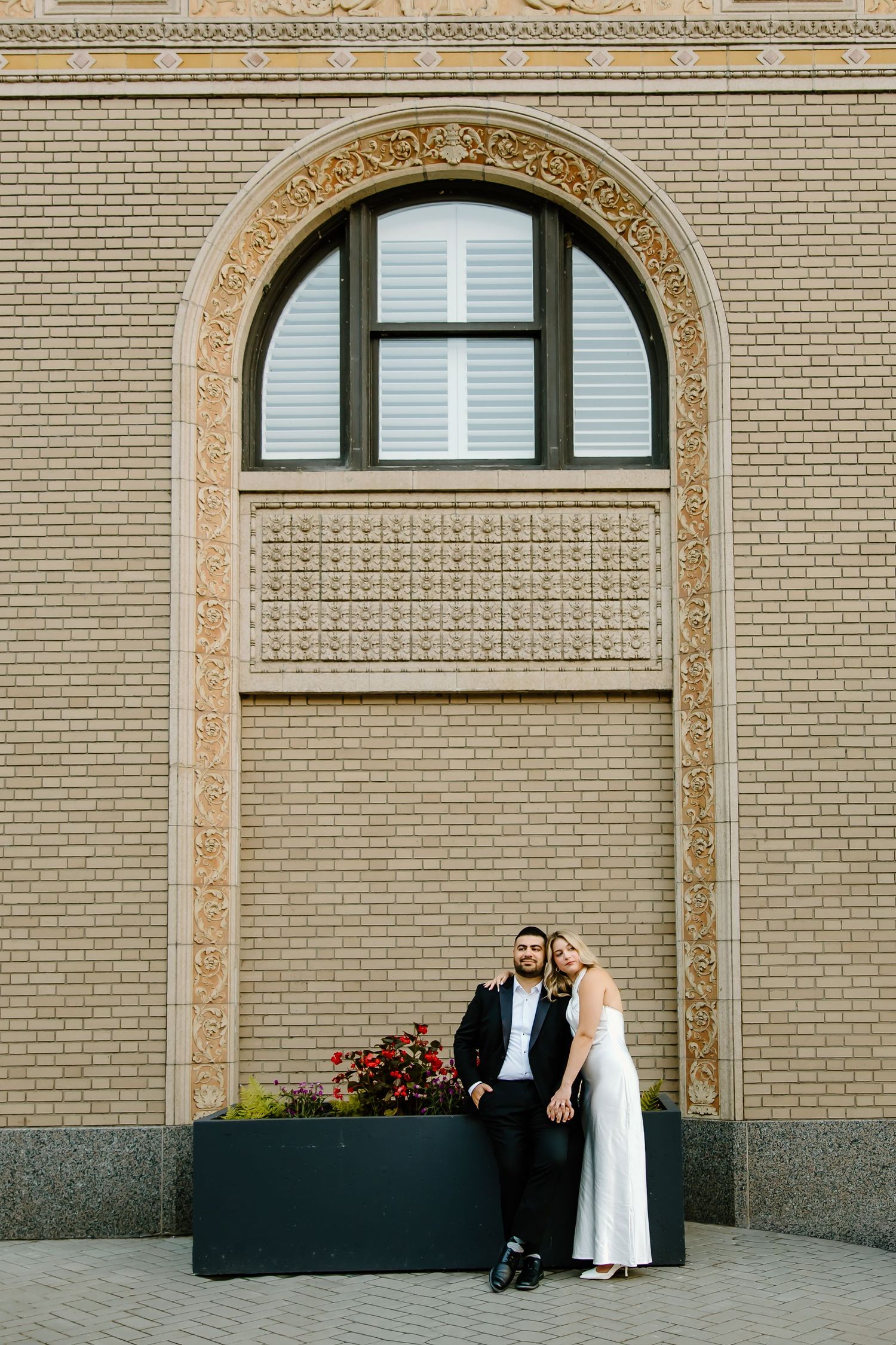 Couple in formal wear sitting beneath decorative arches on a brick wall.