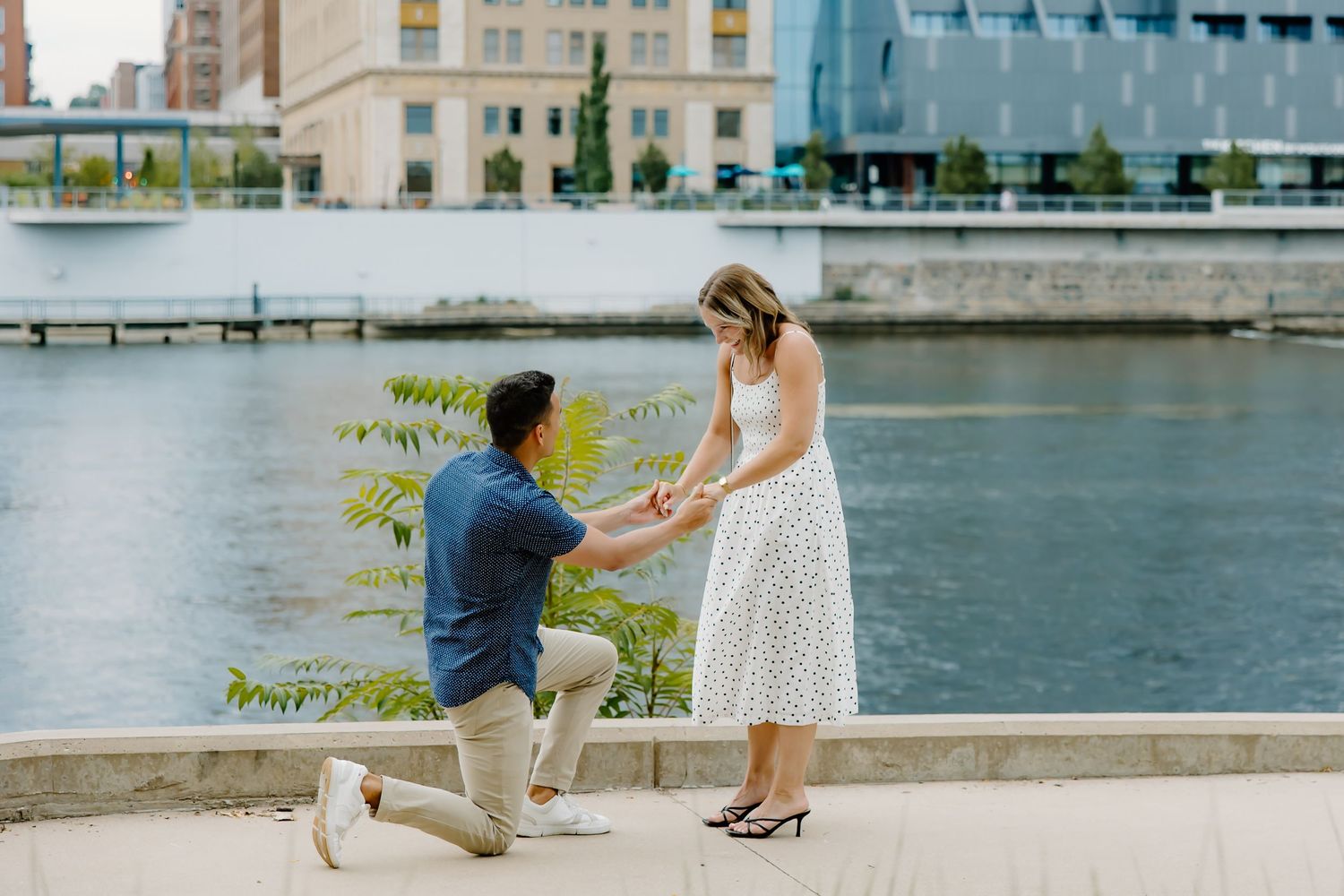 A couple shares a romantic moment along a waterfront with city buildings in the background.