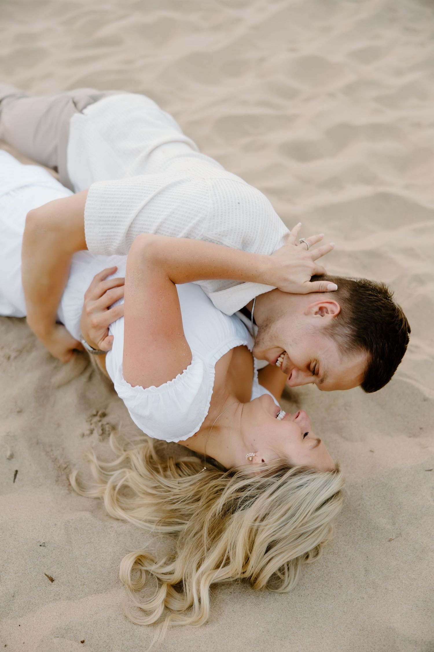 A romantic couple in white outfits embraces while lying on sandy beach.