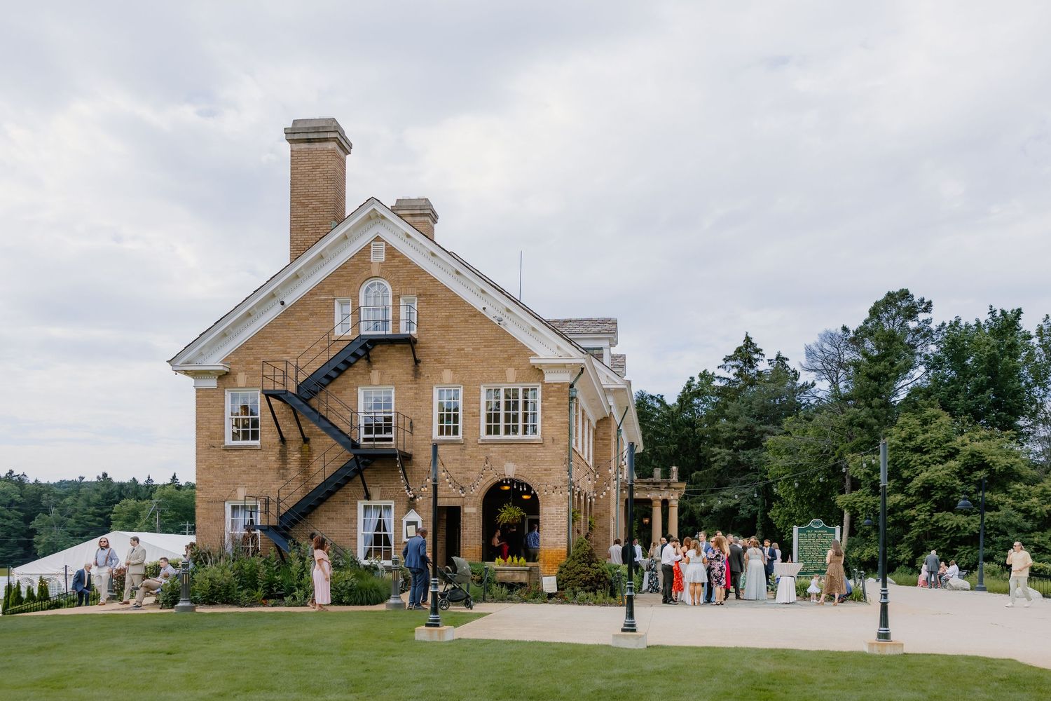 Elegant brick manor house with outdoor wedding guests gathered on manicured lawn surrounded by lush trees.