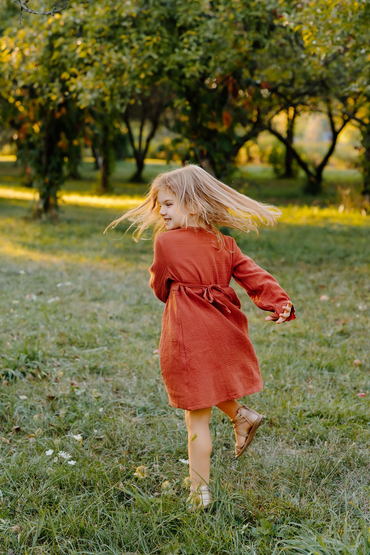 A young child in a rust-colored dress twirls and dances in a sunlit orchard.