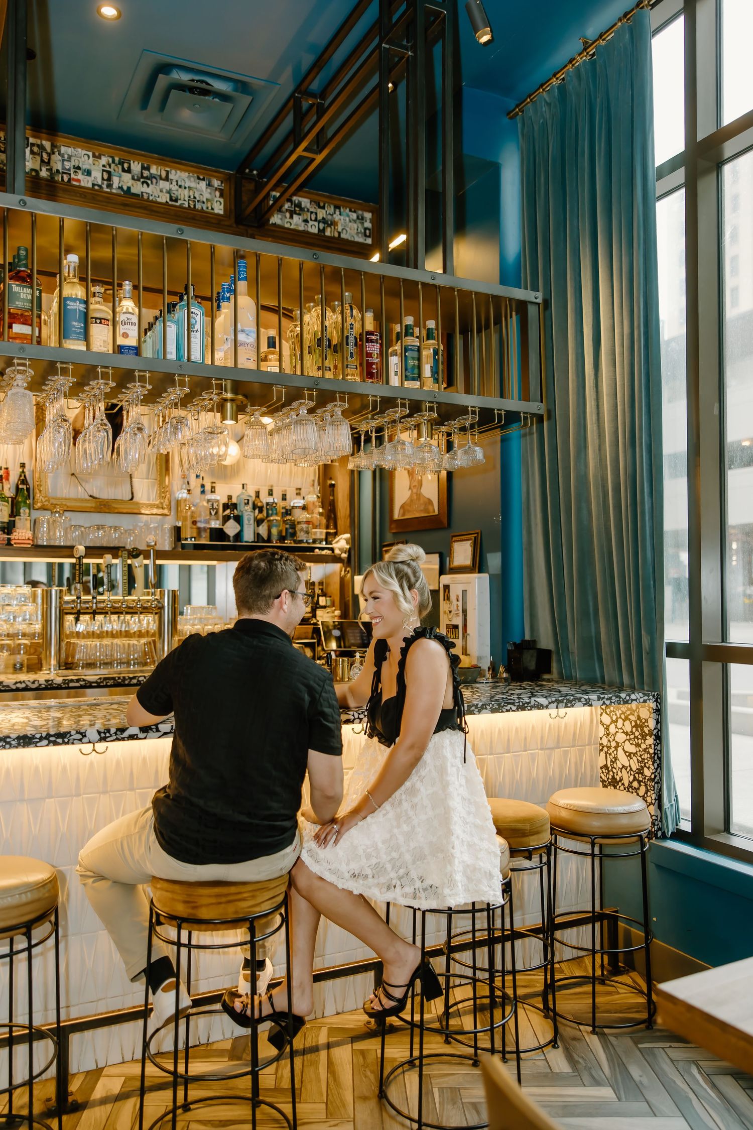Upscale bar interior featuring teal walls, hanging glassware, and modern bar seating.