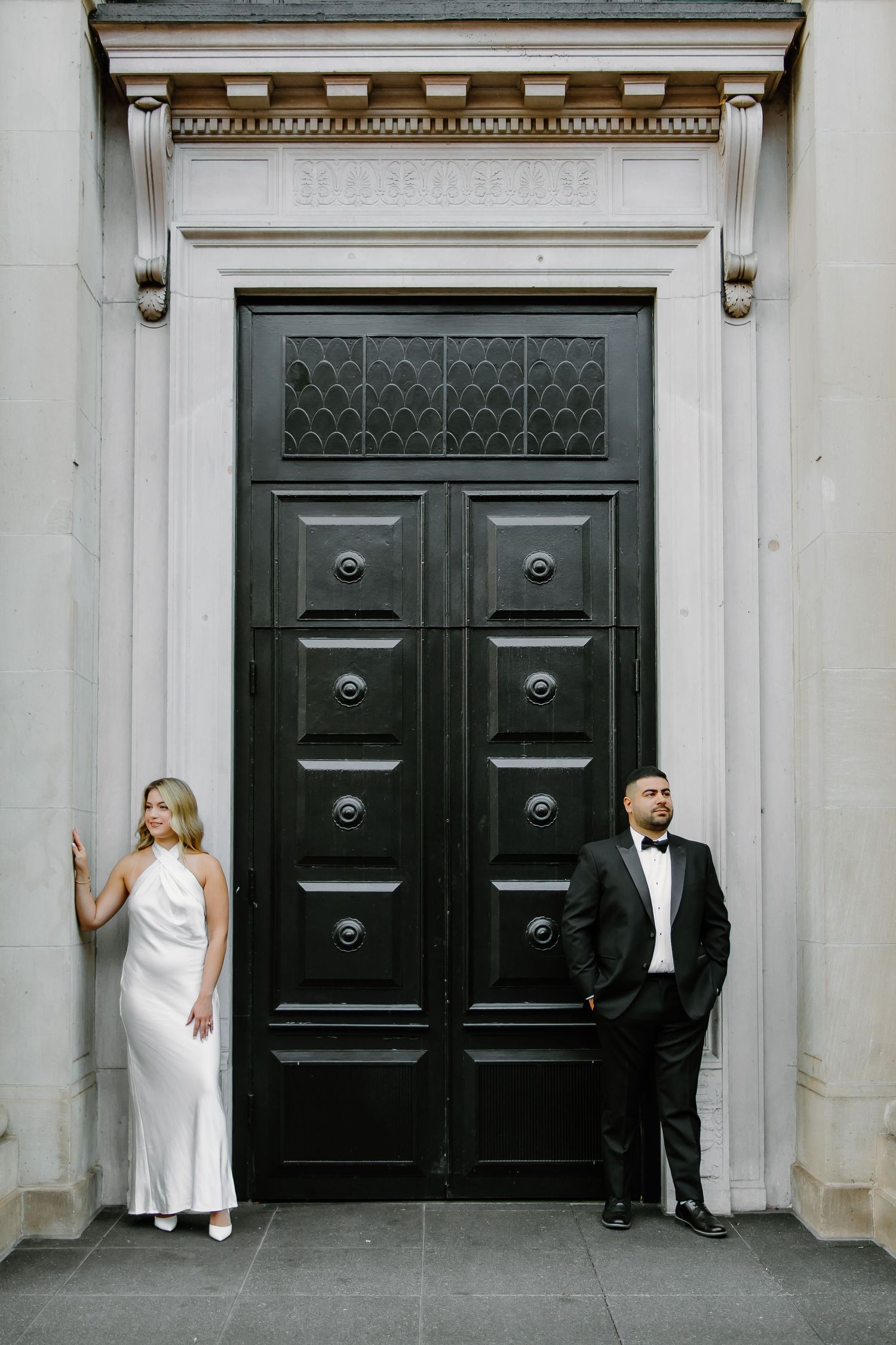 Stylish couple in formal attire poses against large ornate black door.