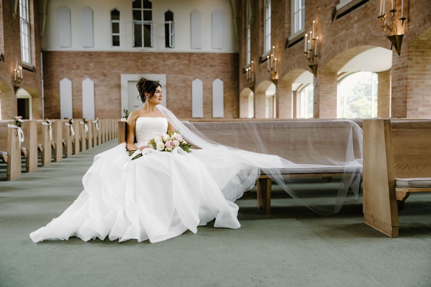 Bride in voluminous white gown sitting gracefully on church pew with dramatic lighting.