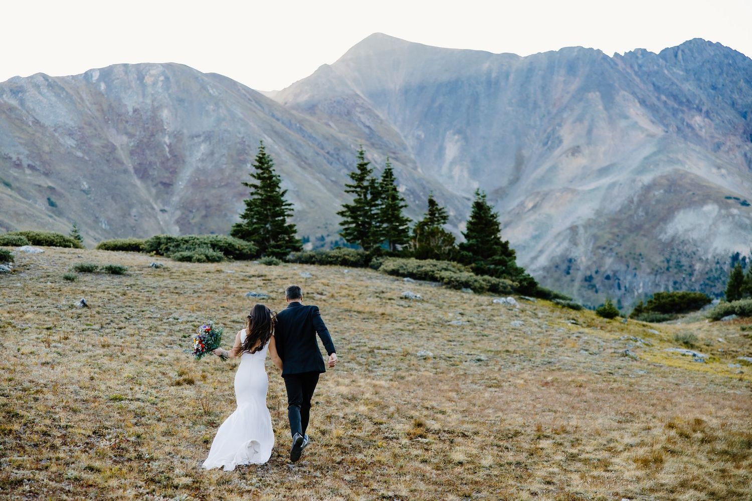 Wedding couple strolls along scenic mountain path surrounded by evergreen trees and rocky peaks.