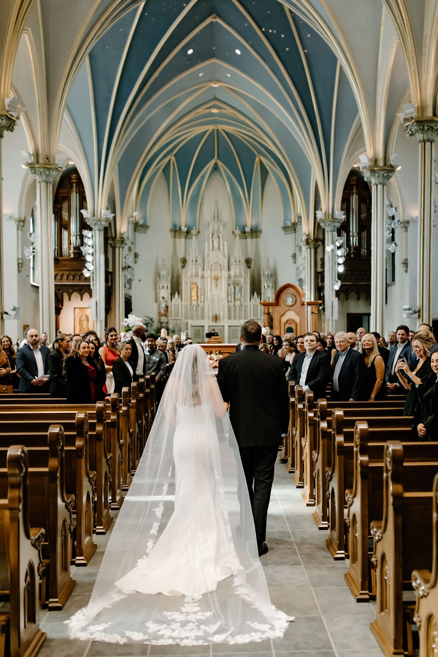 A wedding ceremony processional in a grand cathedral with vaulted blue ceilings and wooden pews.