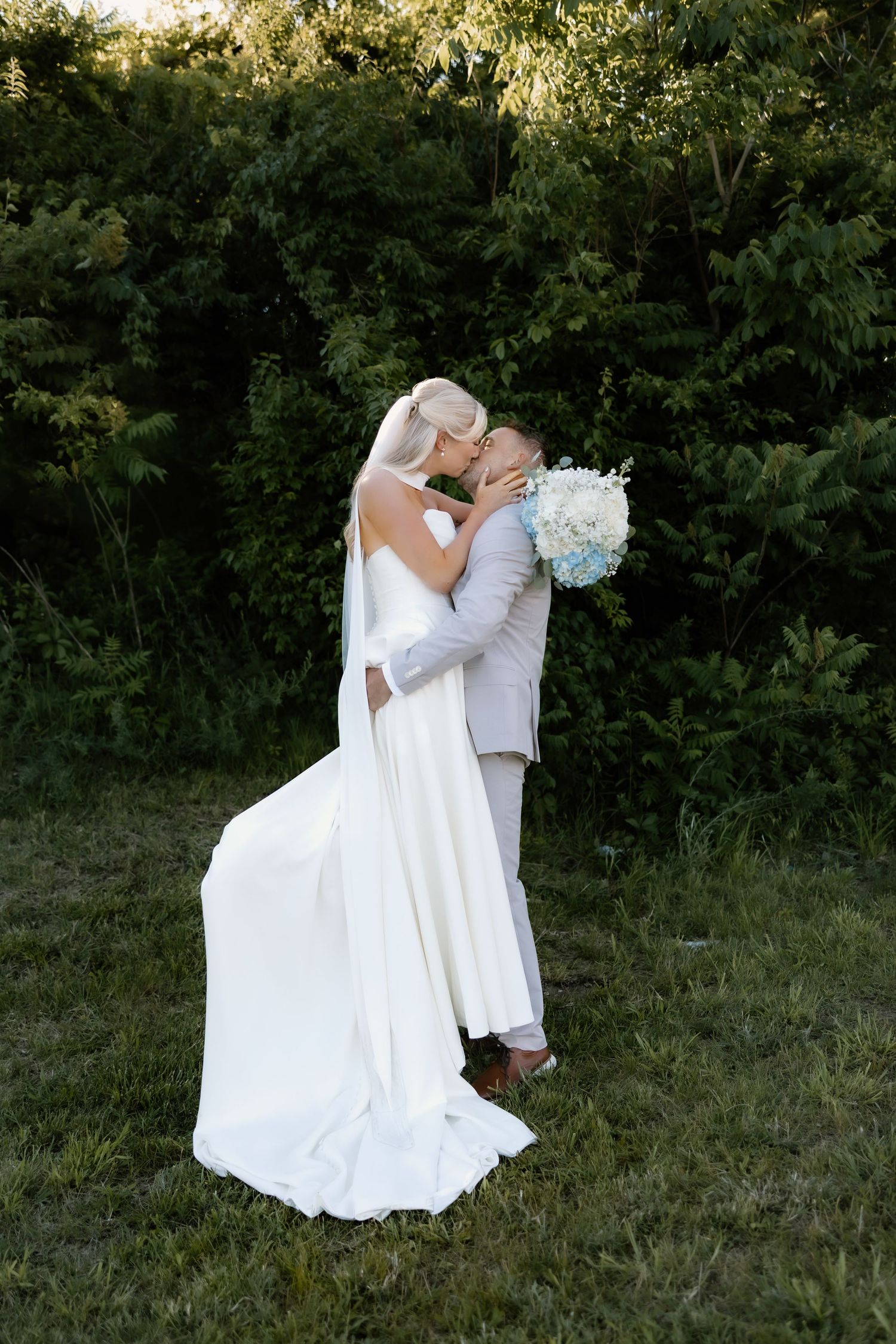 Bride and groom sharing a romantic kiss outdoors, bride in white gown holding blue bouquet, lush green trees background.