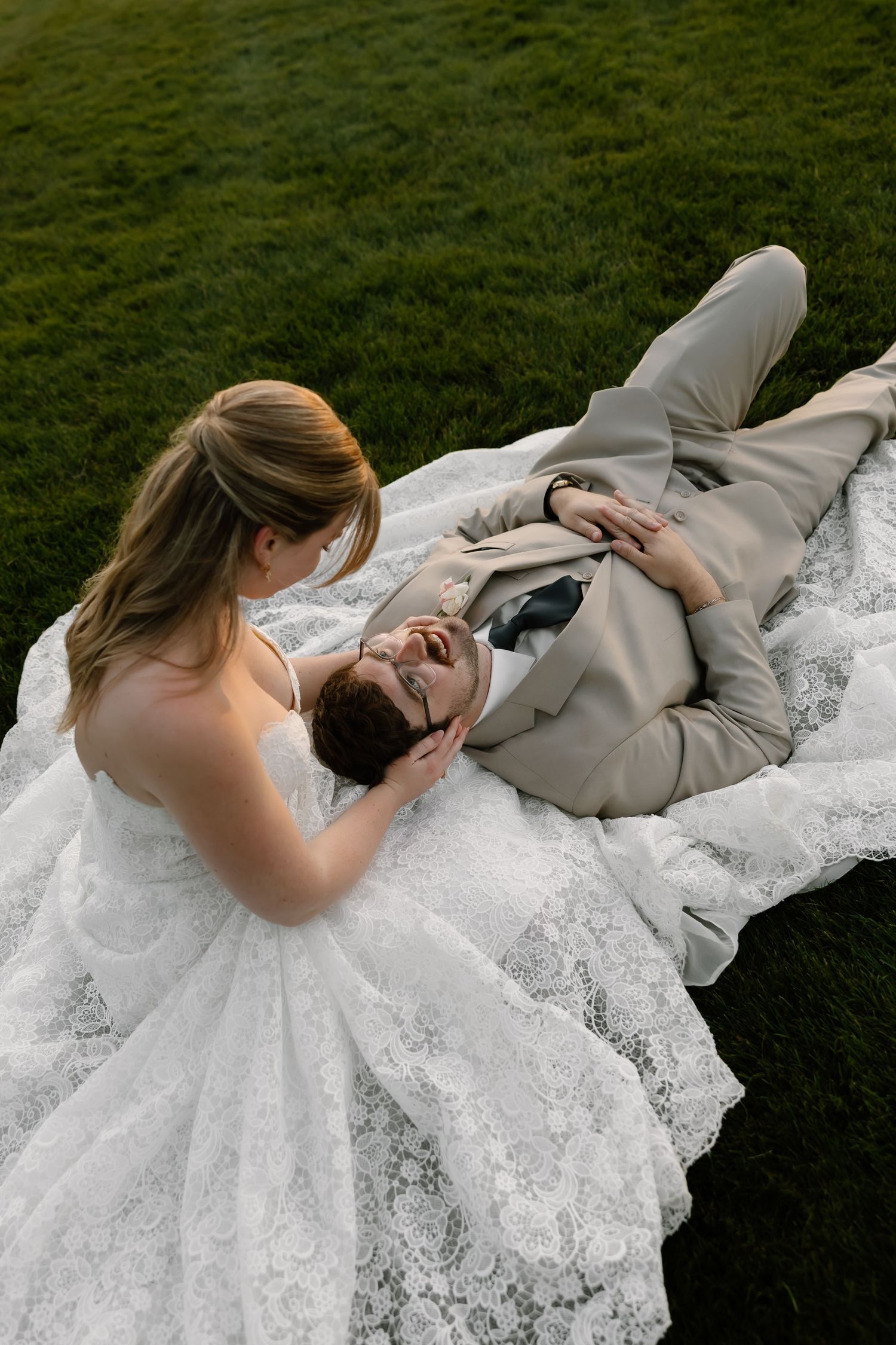 Bride in white lace gown sits beside groom in tan suit lying on green grass, sharing a joyful wedding moment.