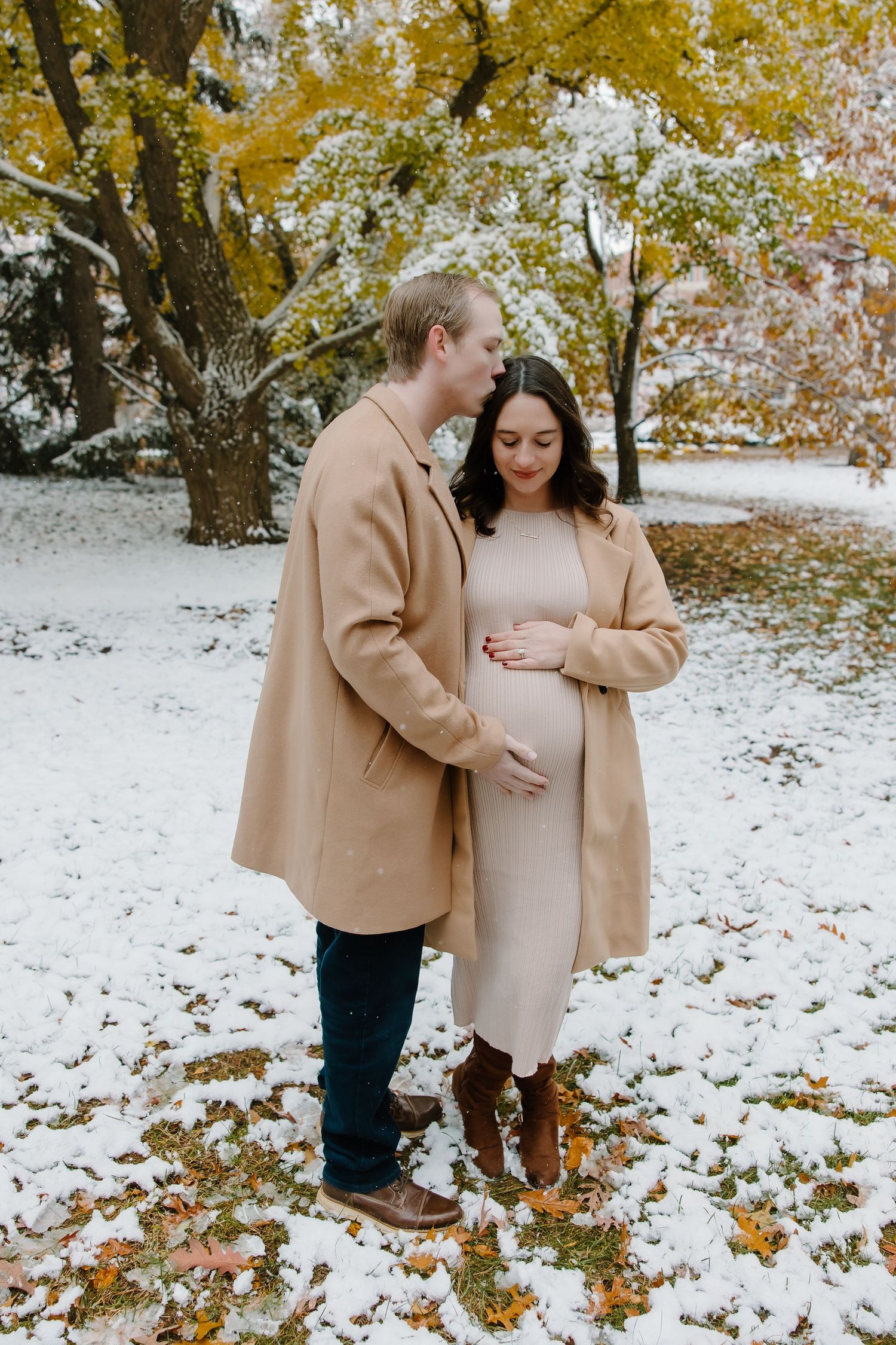 Couple in tan coats standing together on snow-covered ground with autumn leaves and trees in background.