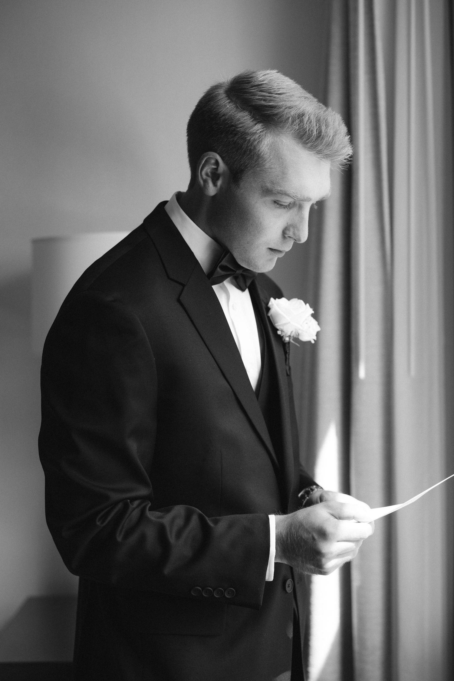 Groom in classic black tuxedo reads a note by window light in dramatic black and white portrait.