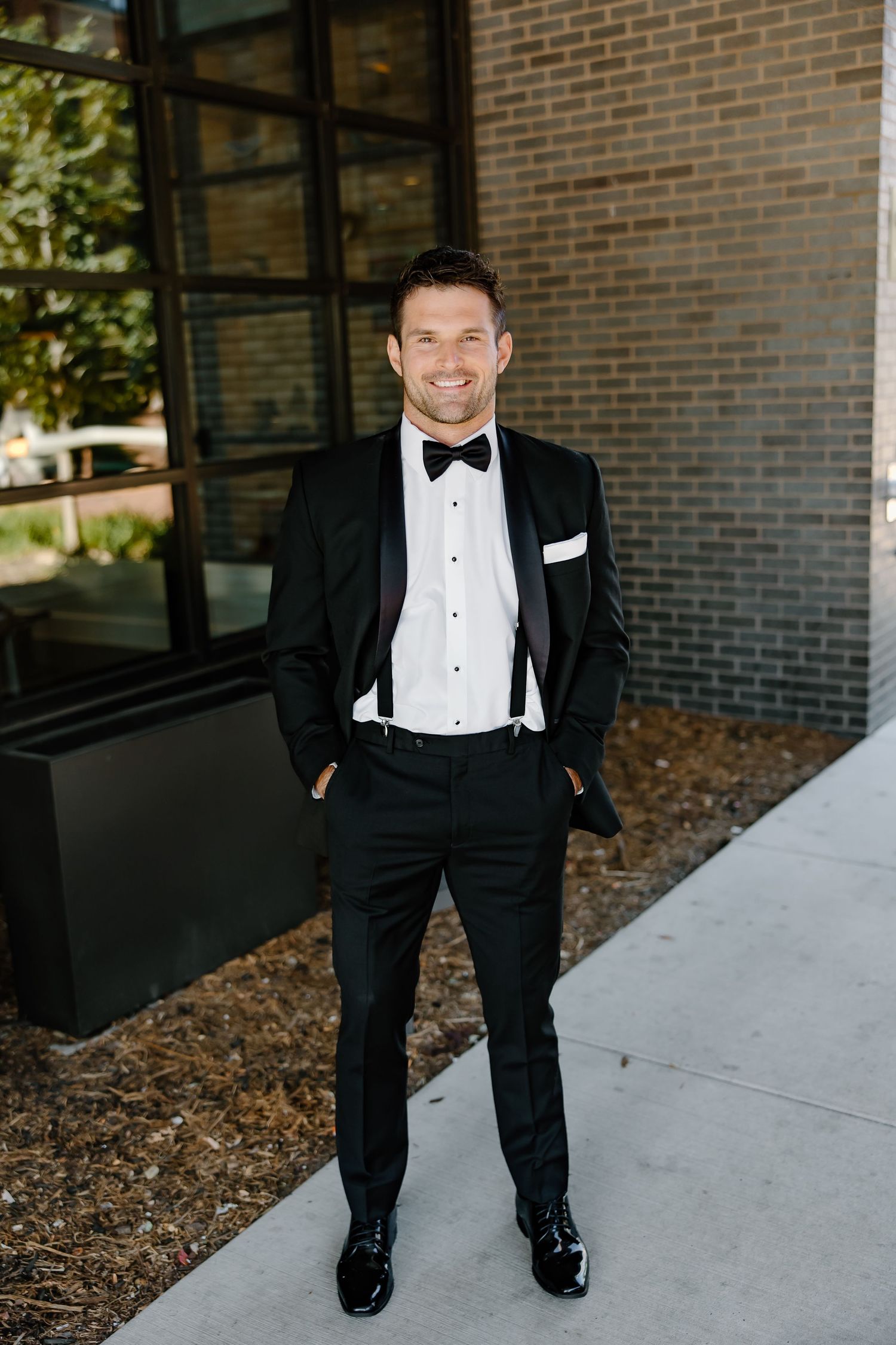 A groom in a black tuxedo and bowtie stands confidently against a modern brick building.