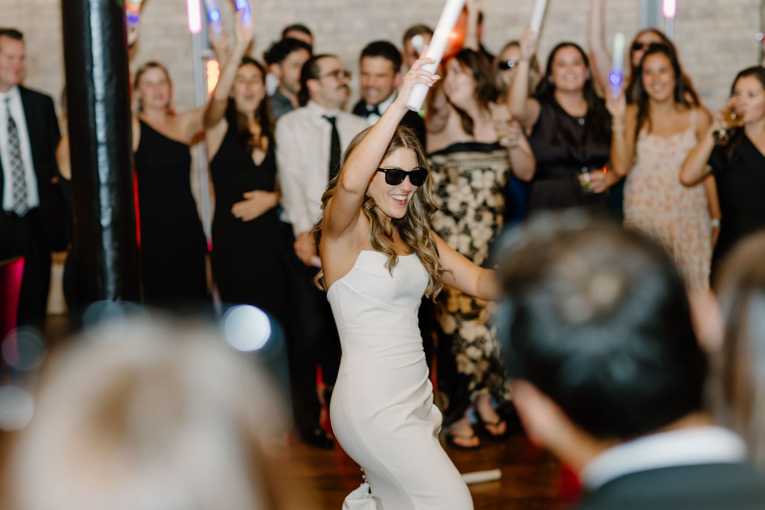Wedding guests dance and celebrate on a crowded dance floor at an indoor reception venue with dim lighting.