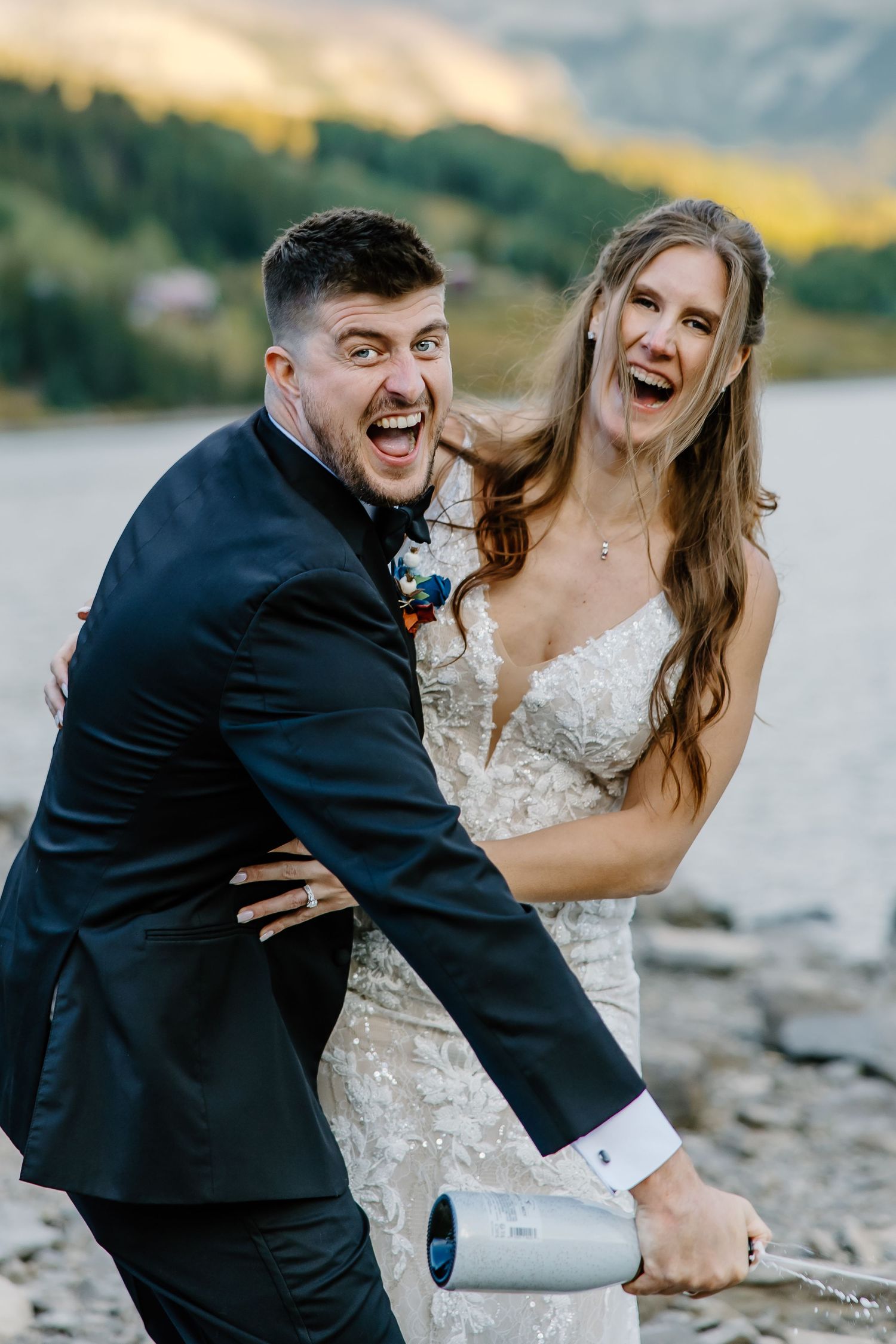 Playful wedding couple laughs while popping champagne by a scenic mountain lake.