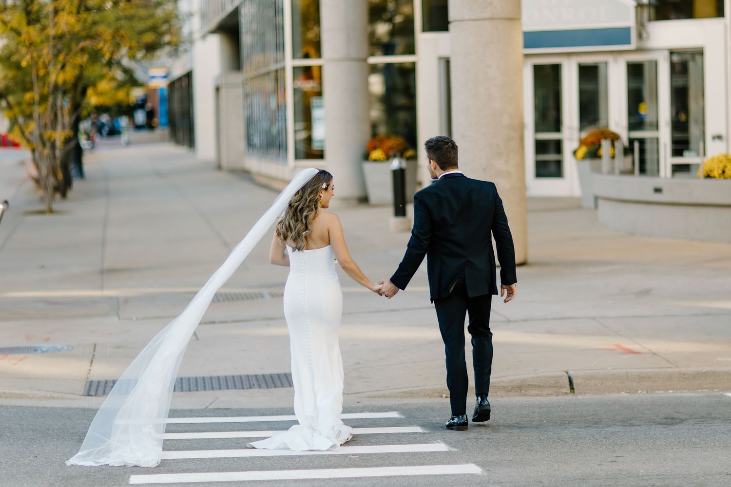Bride and groom walking together on urban sidewalk with flowing veil trailing behind.