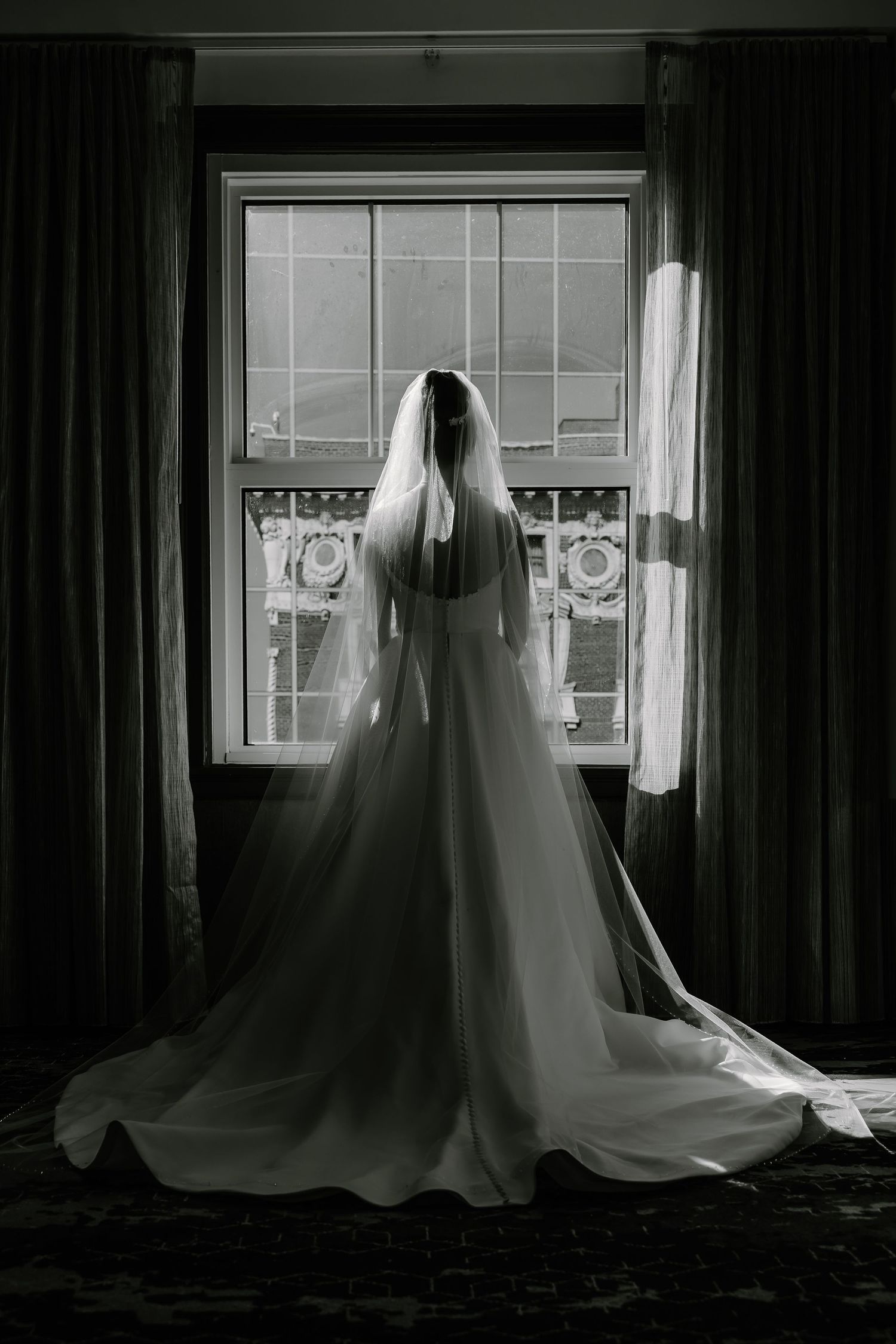 Silhouette of bride in flowing dress and veil against bright window.