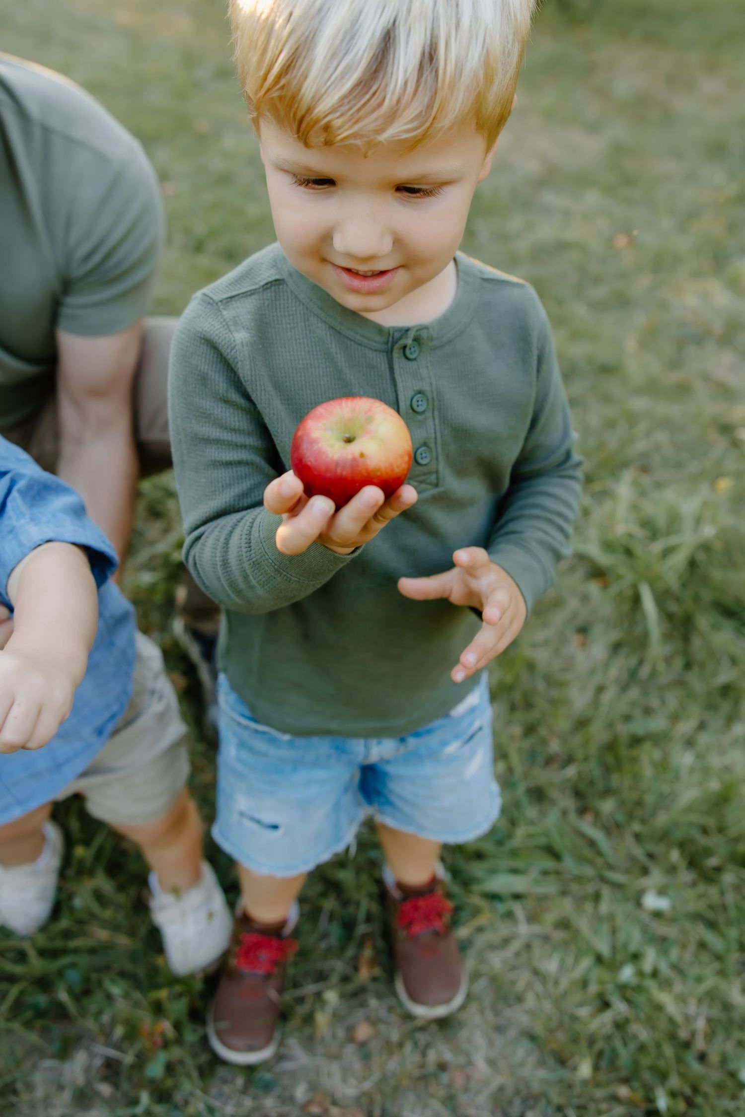 Child in green sweater holds a bright red apple while standing in grass outdoors.