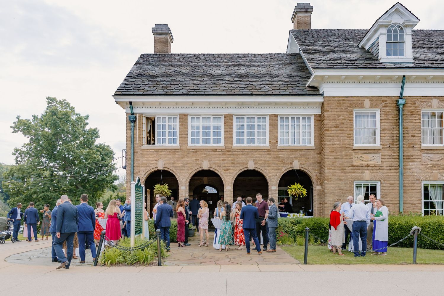 Wedding guests gather outside an elegant brick venue with arched colonnade for an outdoor reception ceremony.