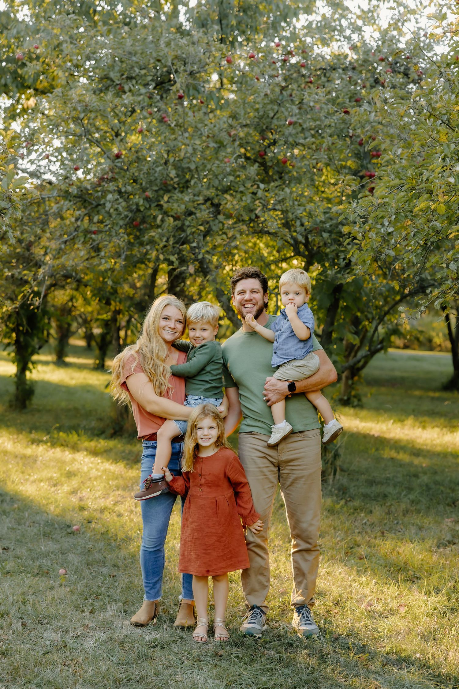 A family of five poses together in an orchard during golden hour with trees in the background.
