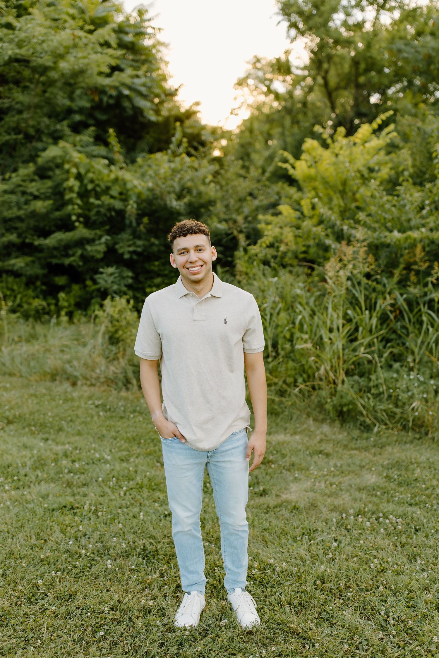 A person wearing a light colored polo shirt and jeans poses in a grassy outdoor setting during golden hour.