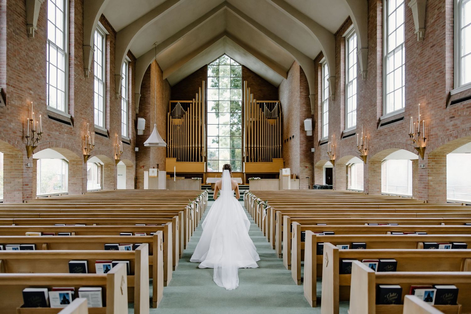 Long view down church aisle with wooden pews and pipe organ, showing flowing white wedding dress.