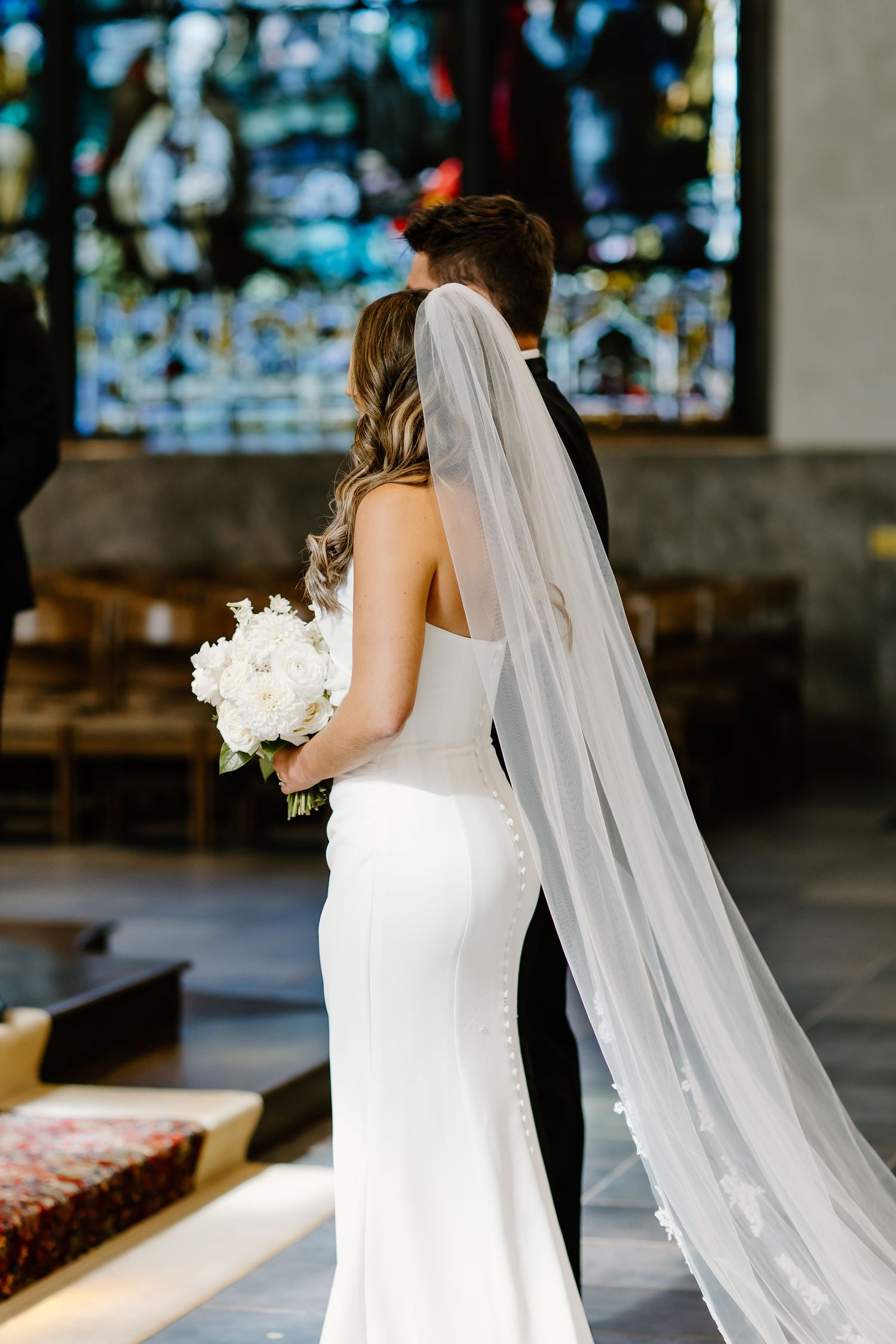 A bride in a fitted white gown and veil stands before stained glass windows.