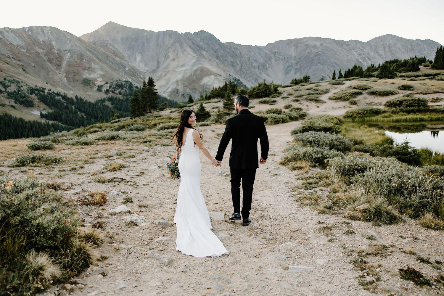 Newlyweds walking along mountain trail with majestic peaks and alpine meadows in the background.