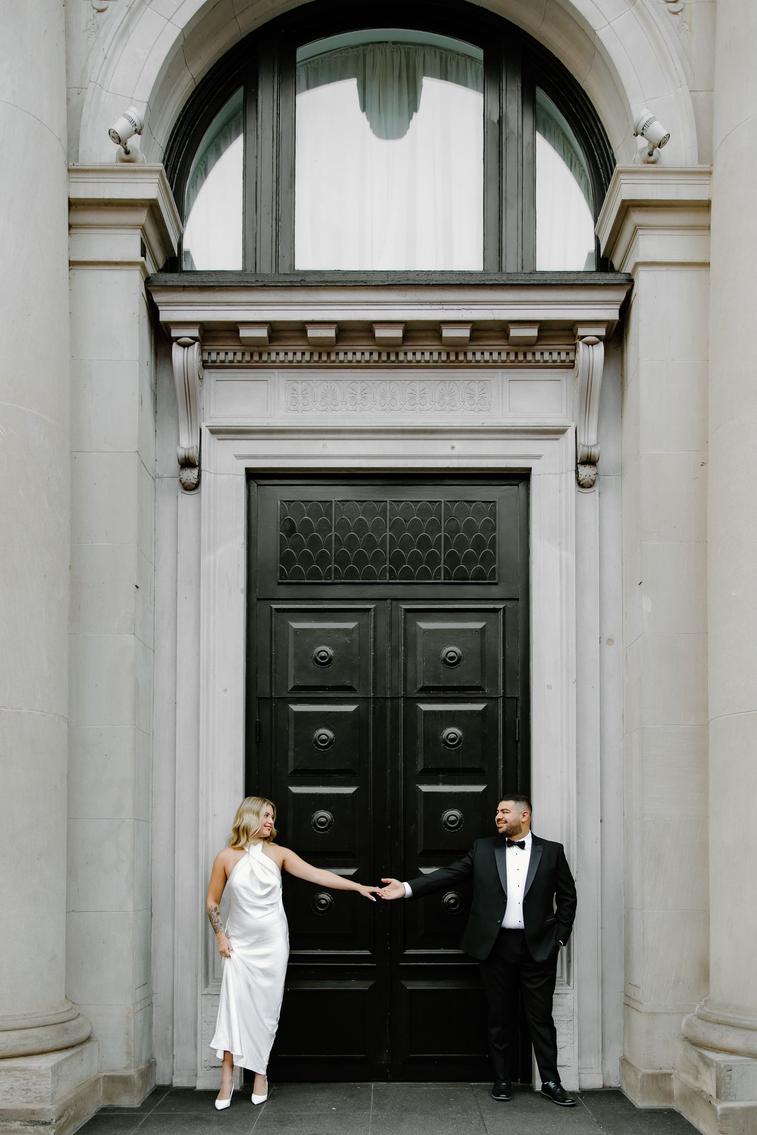 Couple poses elegantly in formal wear by grand black door with arched entrance.