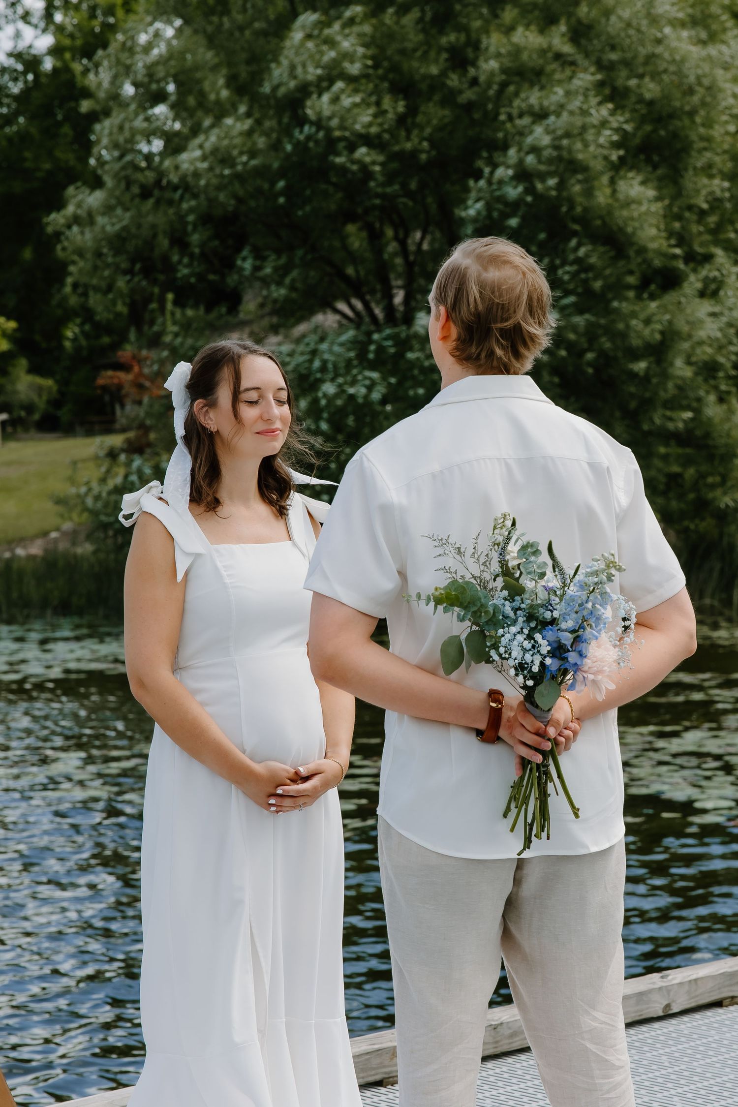 Wedding couple holding bouquets of blue flowers on lakeside dock surrounded by greenery.
