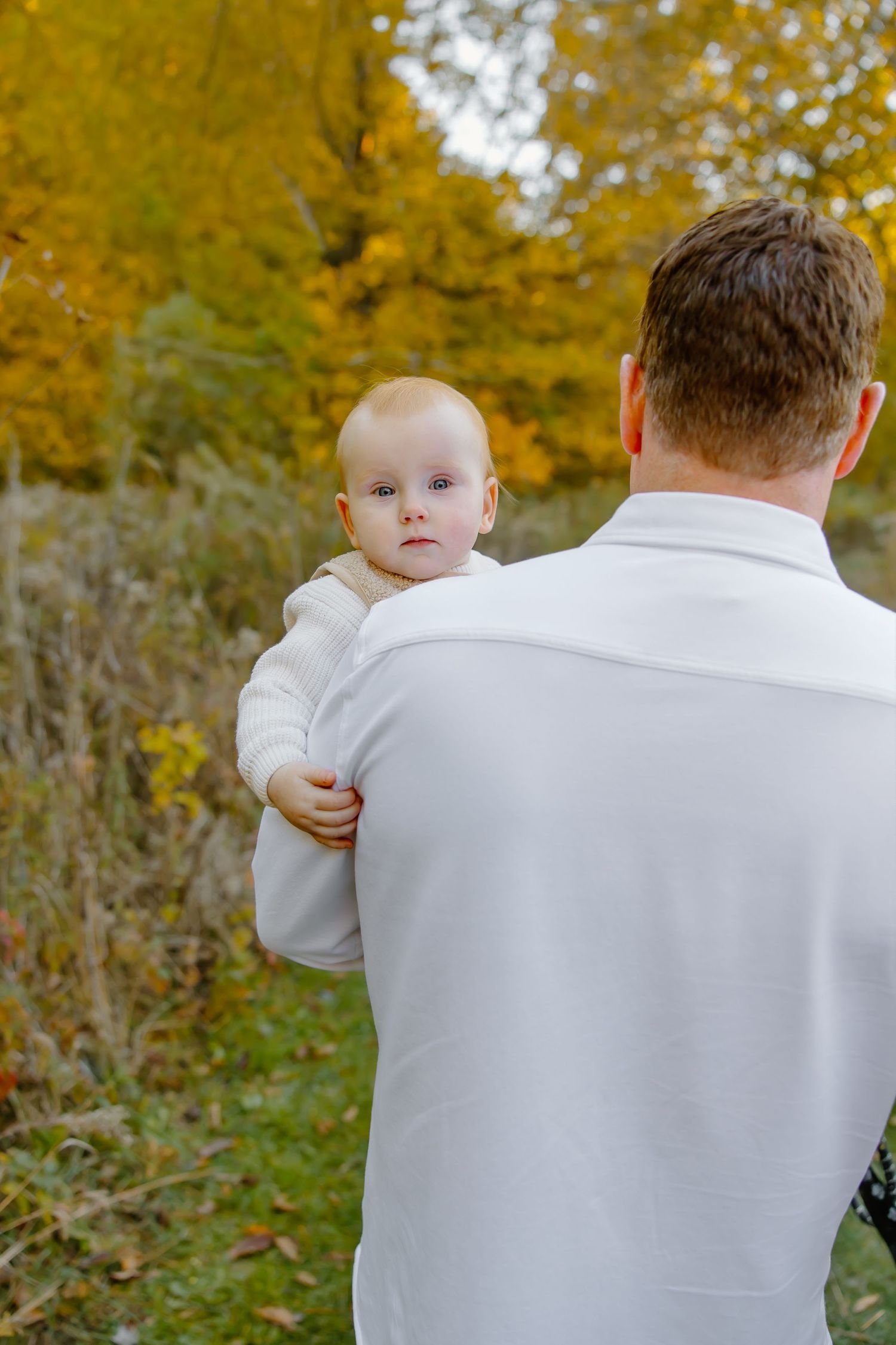 Baby in white sweater being held against autumn background with golden leaves.
