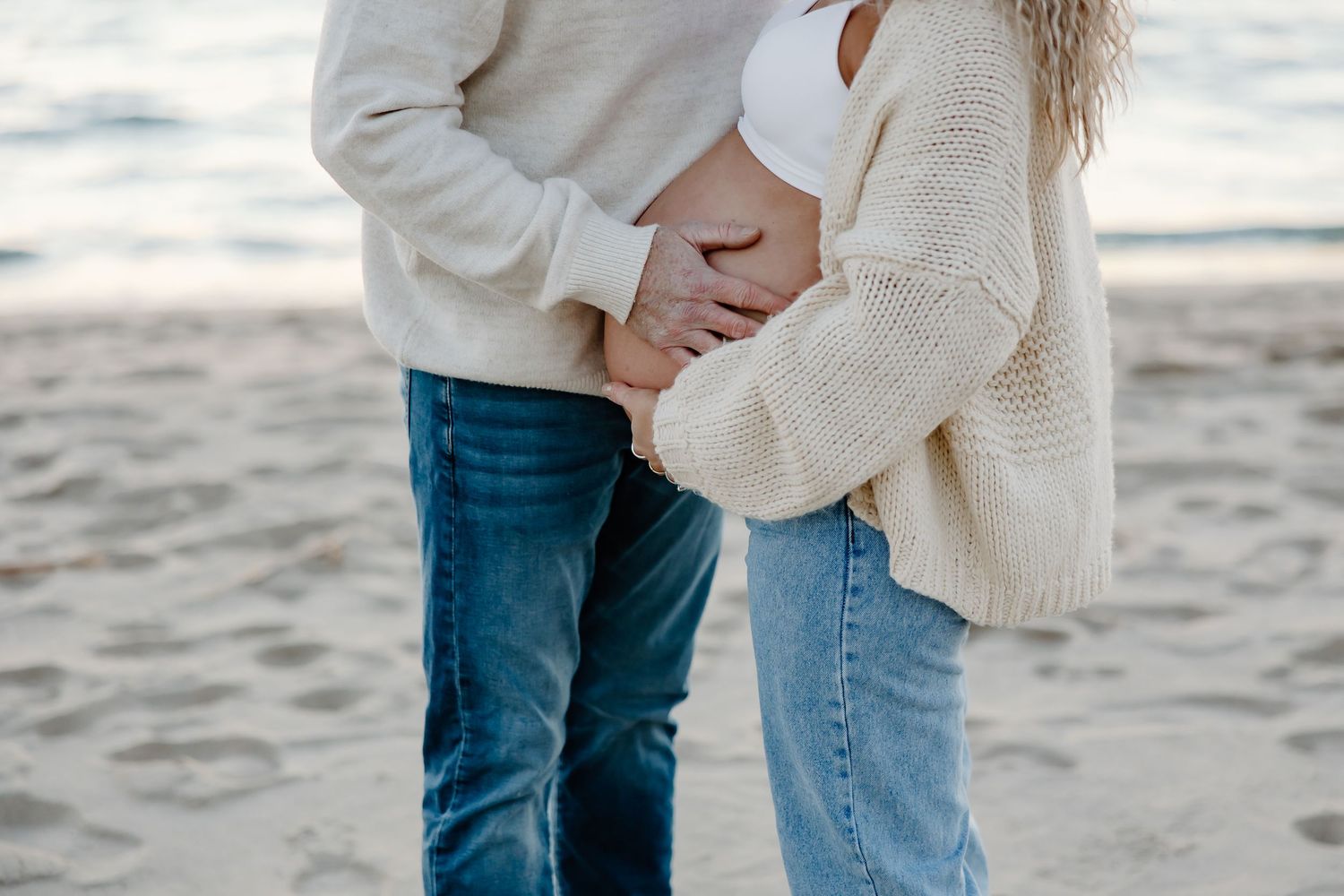Couple wearing white and denim outfits embracing on a sandy beach at sunset.