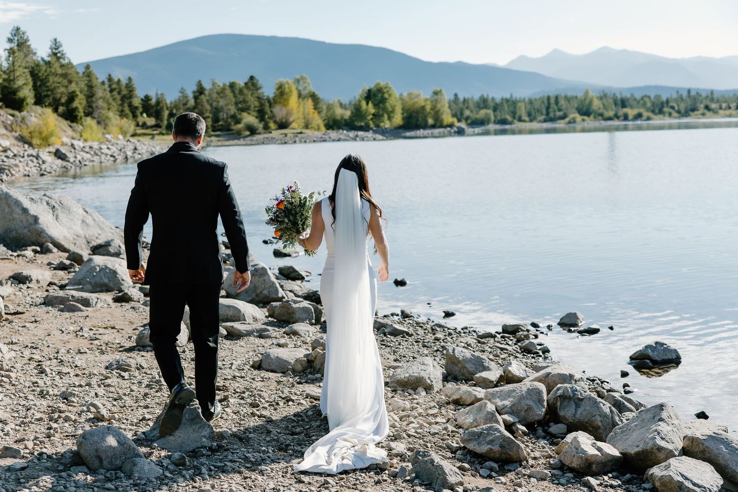 Rocky lakeside landscape with mountains and wedding couple walking along shore.