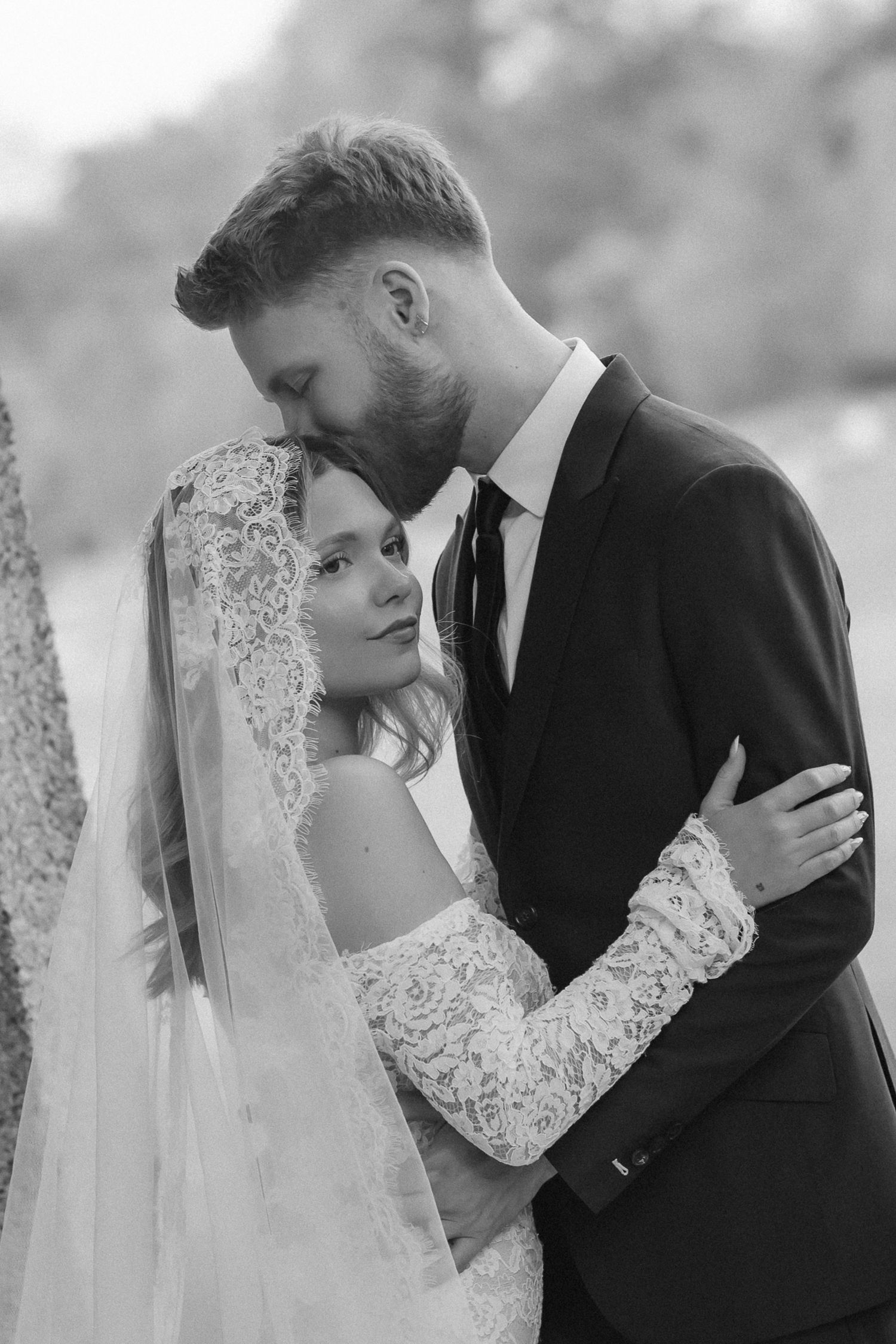 Black and white wedding photo of bride in lace gown and veil embracing groom in dark suit as he kisses her forehead.