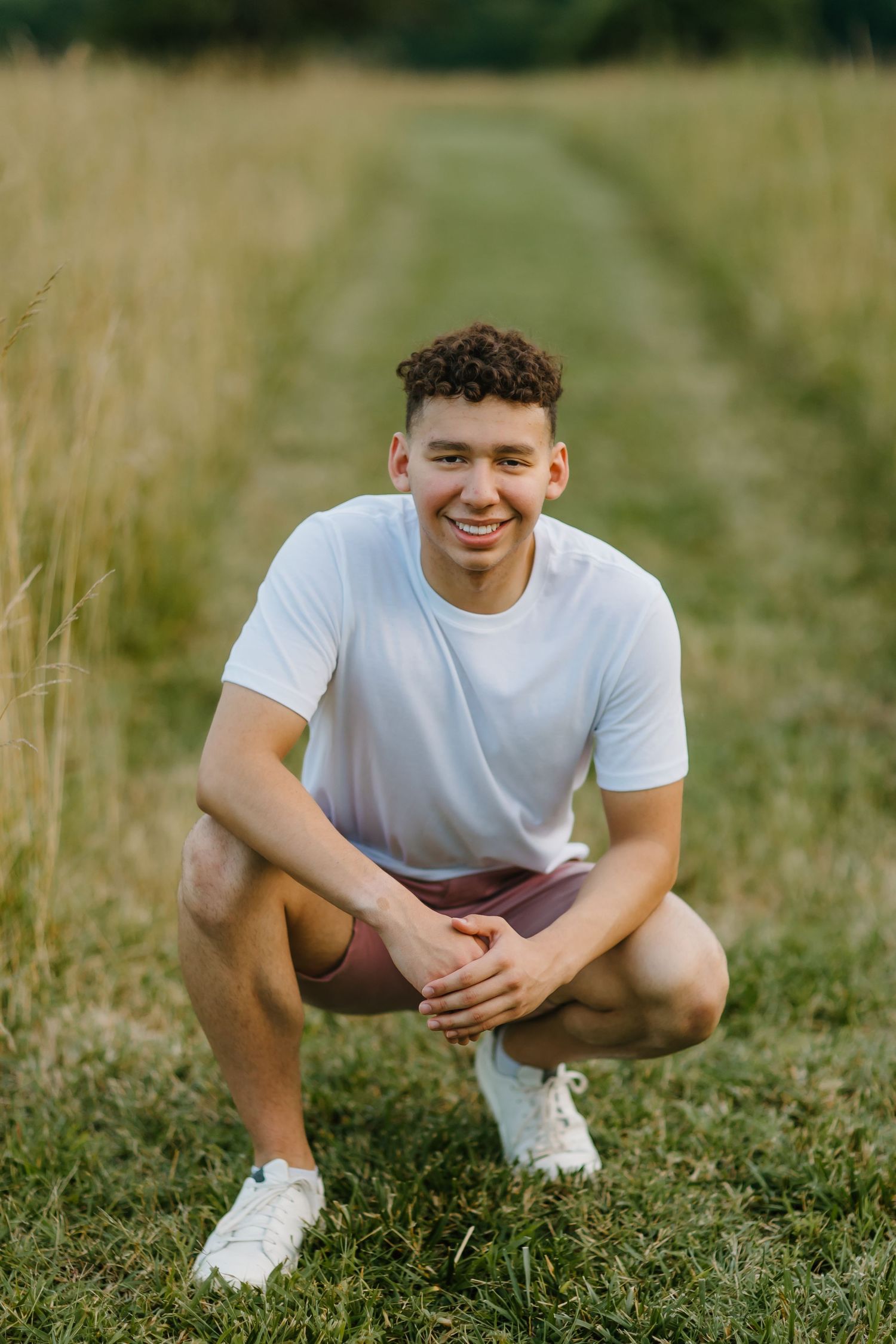 A person in a white t-shirt and shorts crouches in a grassy field on a sunny day.