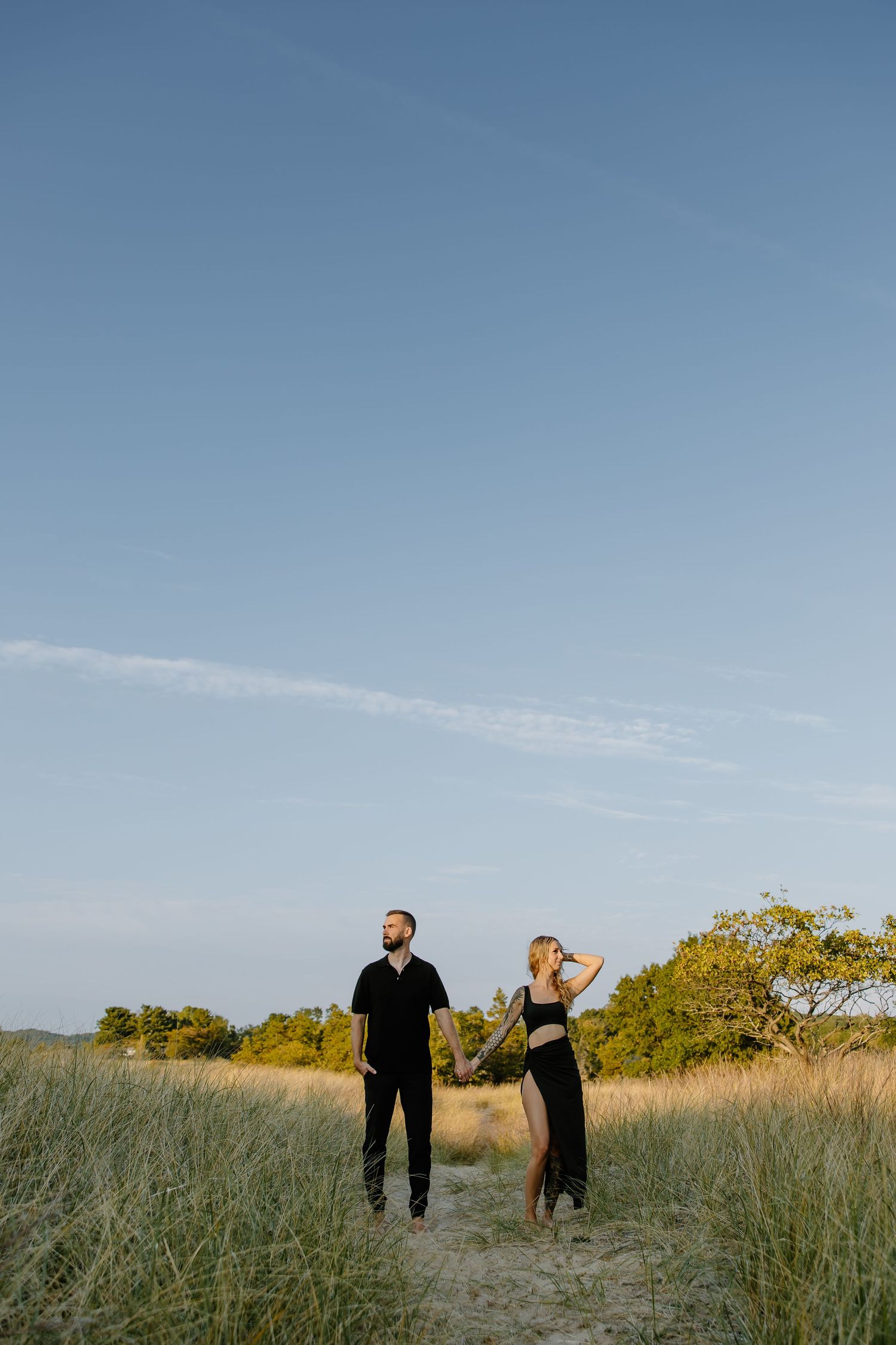 Couple walking through tall grass in open field against blue sky.