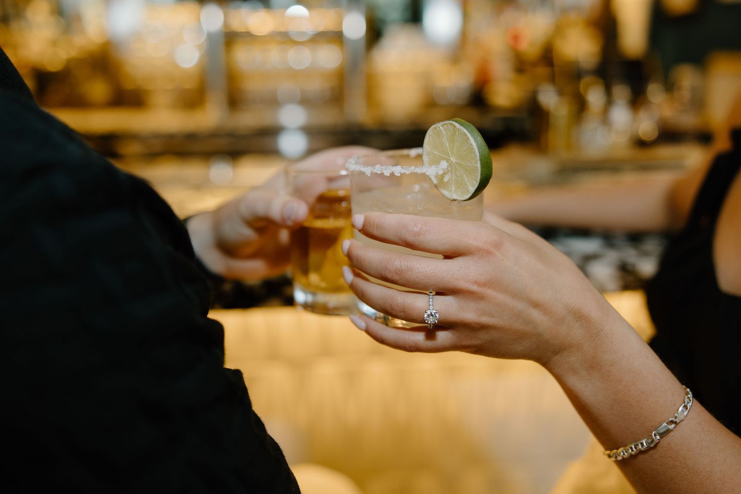 Close-up of hands holding cocktail glasses at a dimly lit bar.