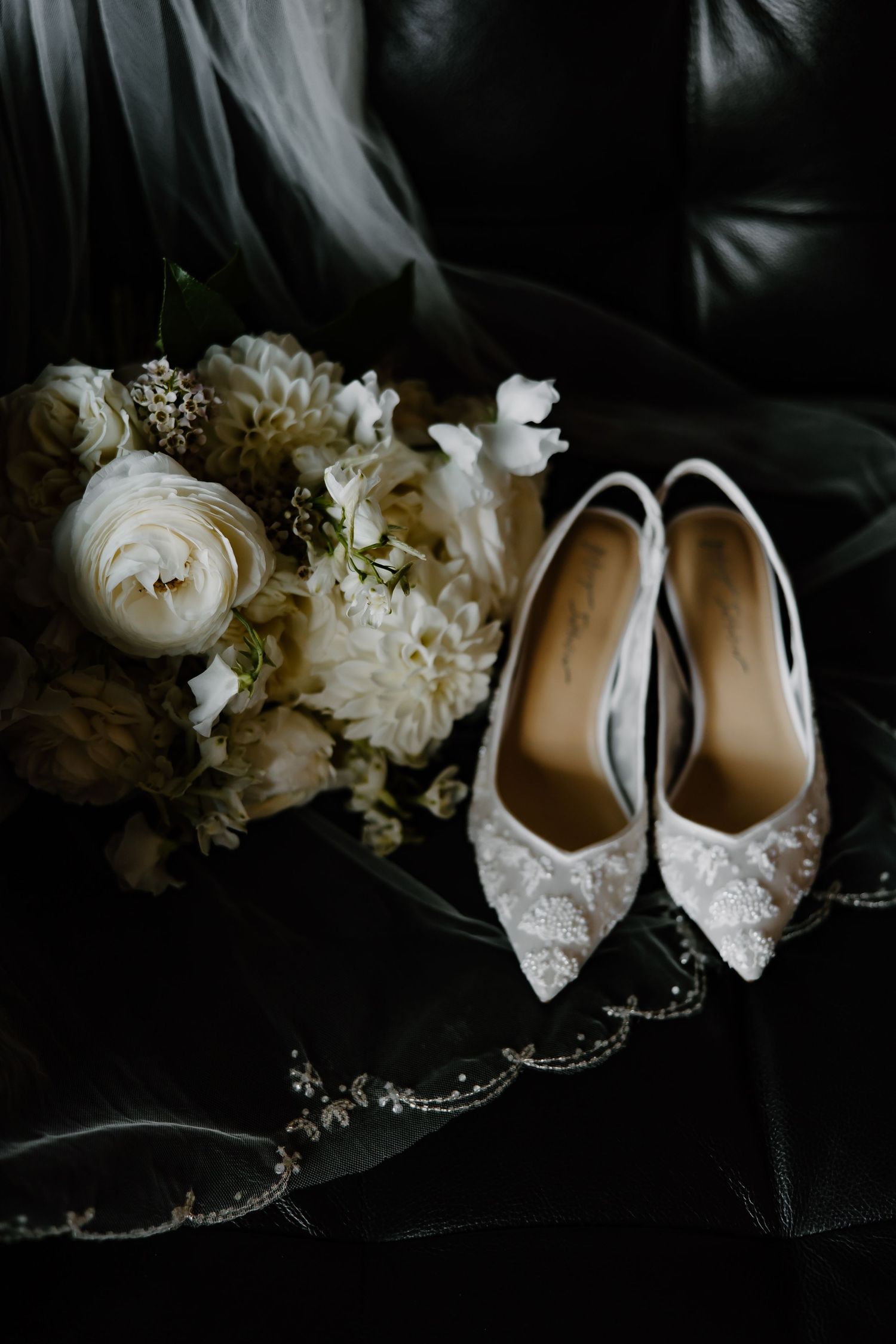 Elegant crystal-embellished bridal shoes next to white roses on dark leather.
