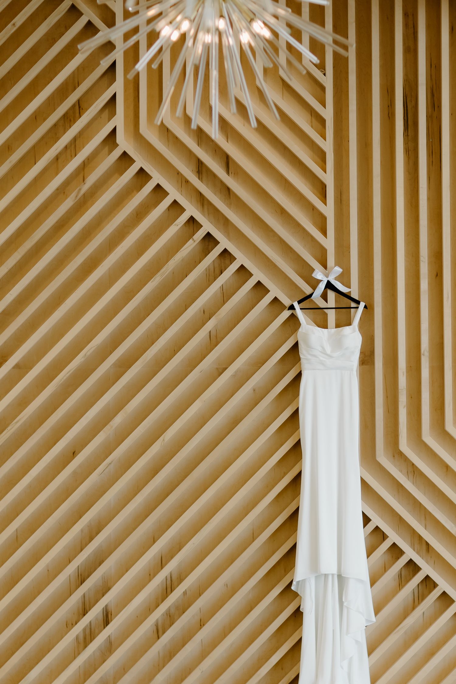 Modern white wedding dress hanging against geometric wooden wall pattern.