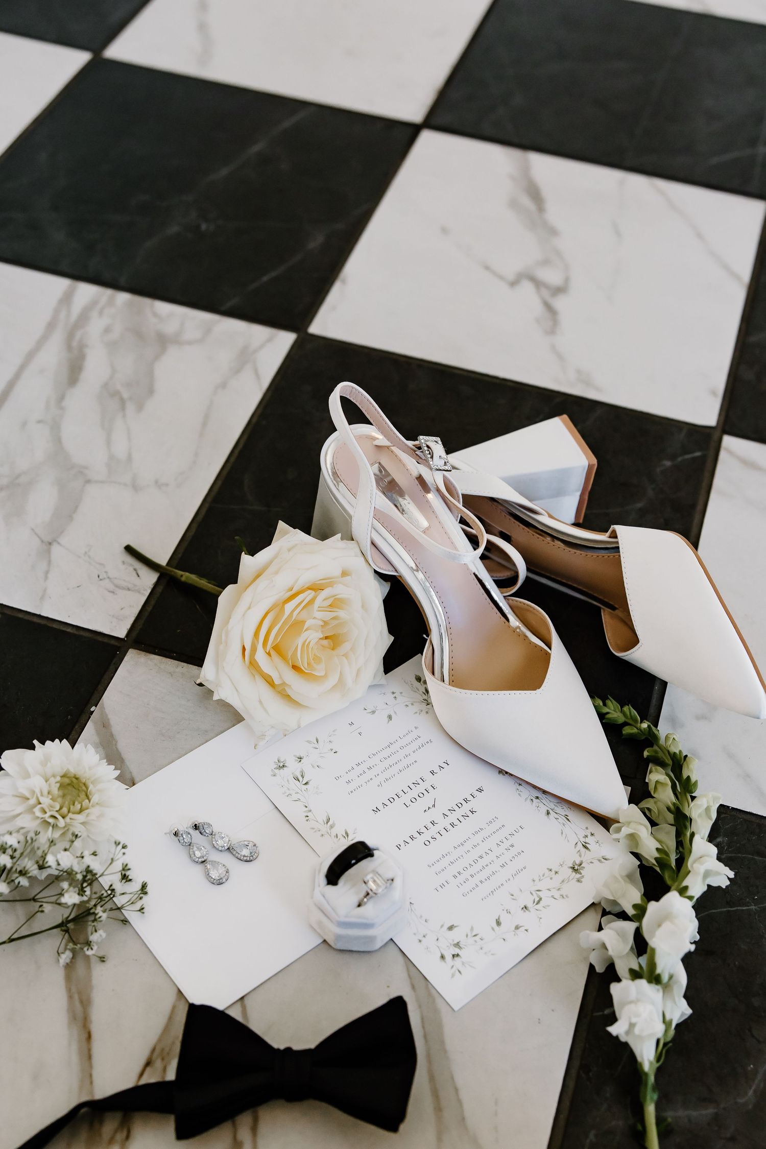 Wedding details on checkered marble floor including white shoes, invitations and roses.