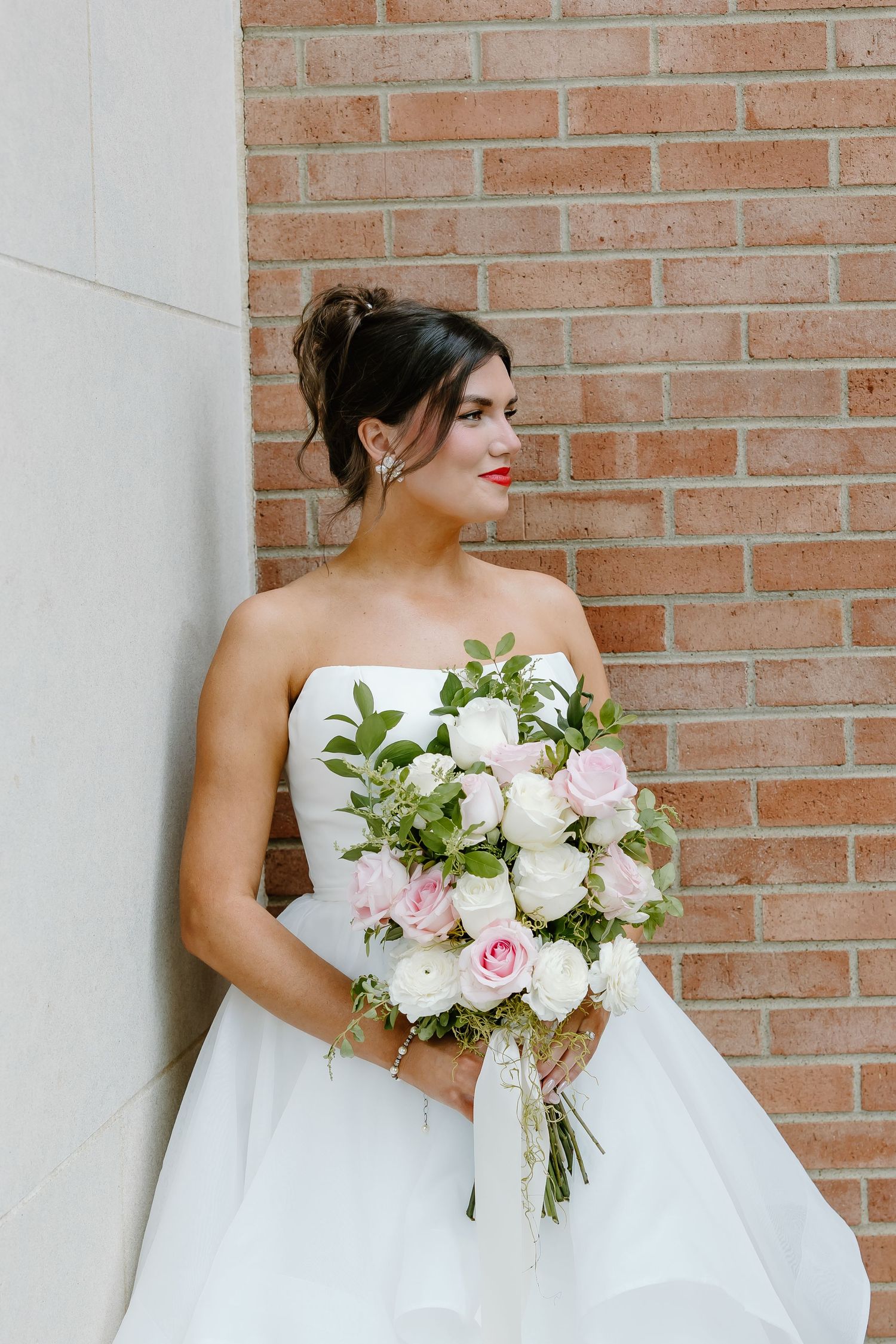 Elegant bride in strapless white gown holding romantic bouquet of pink and white flowers against brick wall.